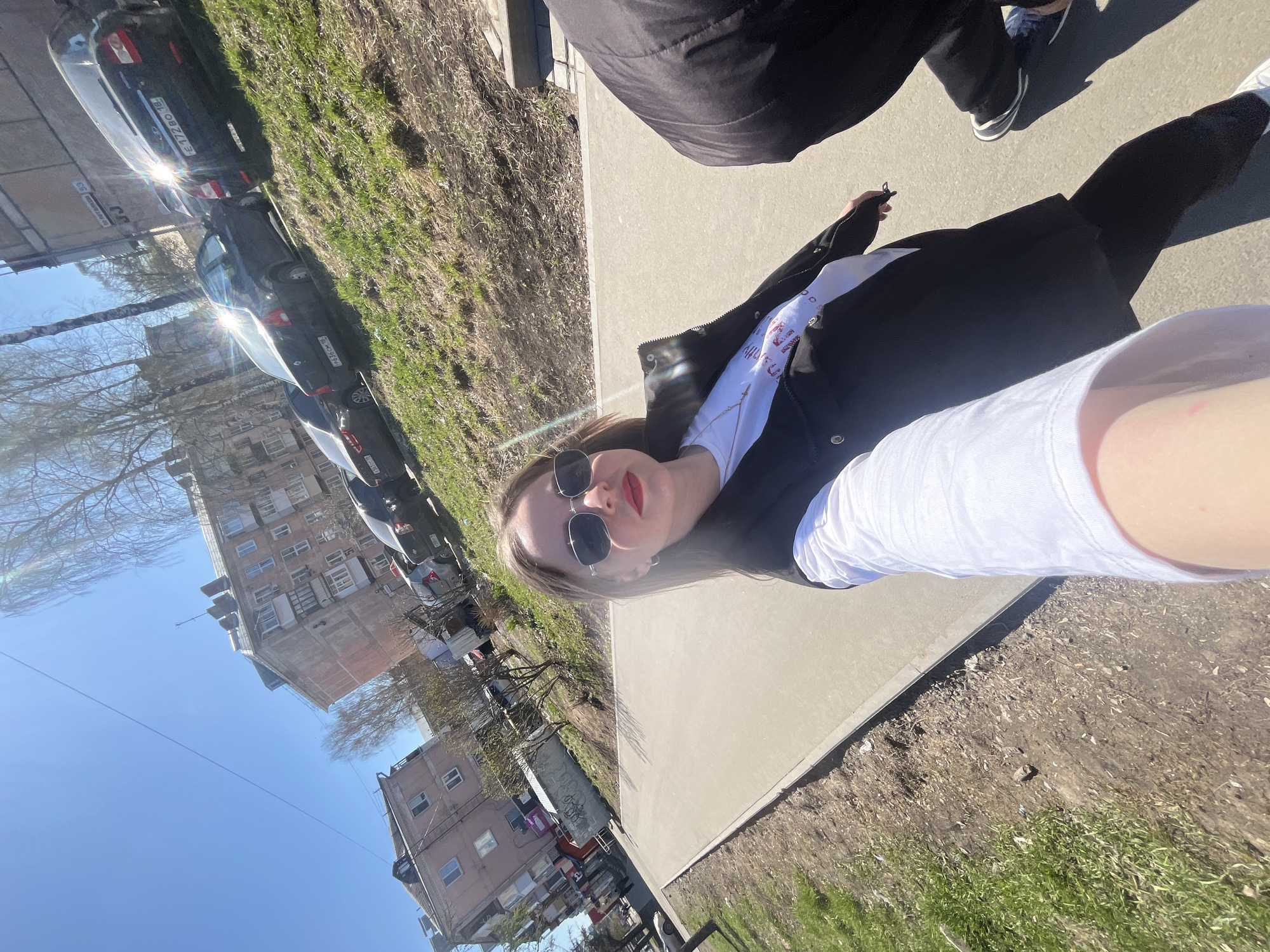 The image appears to be a selfie taken on a city street in Izhevsk, Russia. The buildings and vehicles in the background are consistent with urban landscapes found in the region.
A person, presumably the one taking the selfie, is in the foreground. They are wearing sunglasses, a white top, and a black jacket. Another person wearing black pants and tennis shoes is also partially visible.
The setting is outdoors on a sunny day. There's a concrete sidewalk next to grass and trees. Multiple cars are parked along the street, with visible Russian license plates. The architecture consists of several-story residential buildings.