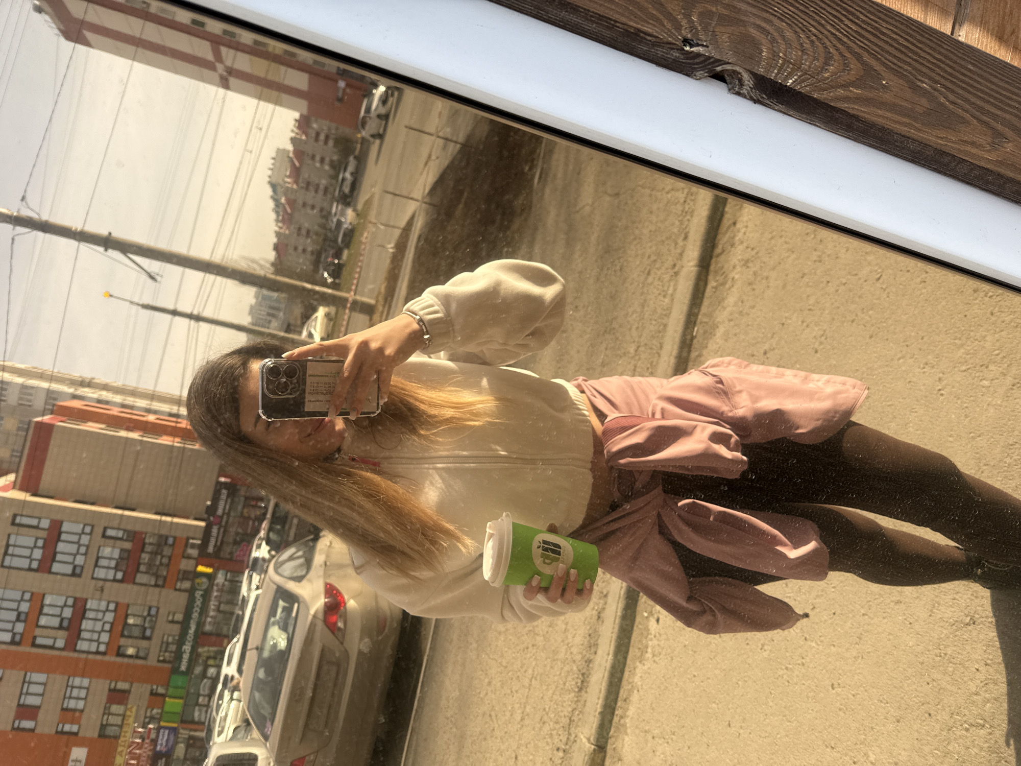 The image appears to be a reflection in a glass window, possibly of a storefront. A young woman is taking a photo of herself in the reflection, holding a green coffee cup in her left hand. She is wearing a white zip-up jacket, a pink skirt tied around her waist, and dark-colored tights. In the reflection, a busy urban street scene is visible with multiple cars parked along the side, multi-story buildings, and power lines overhead. The specific location is not identifiable.