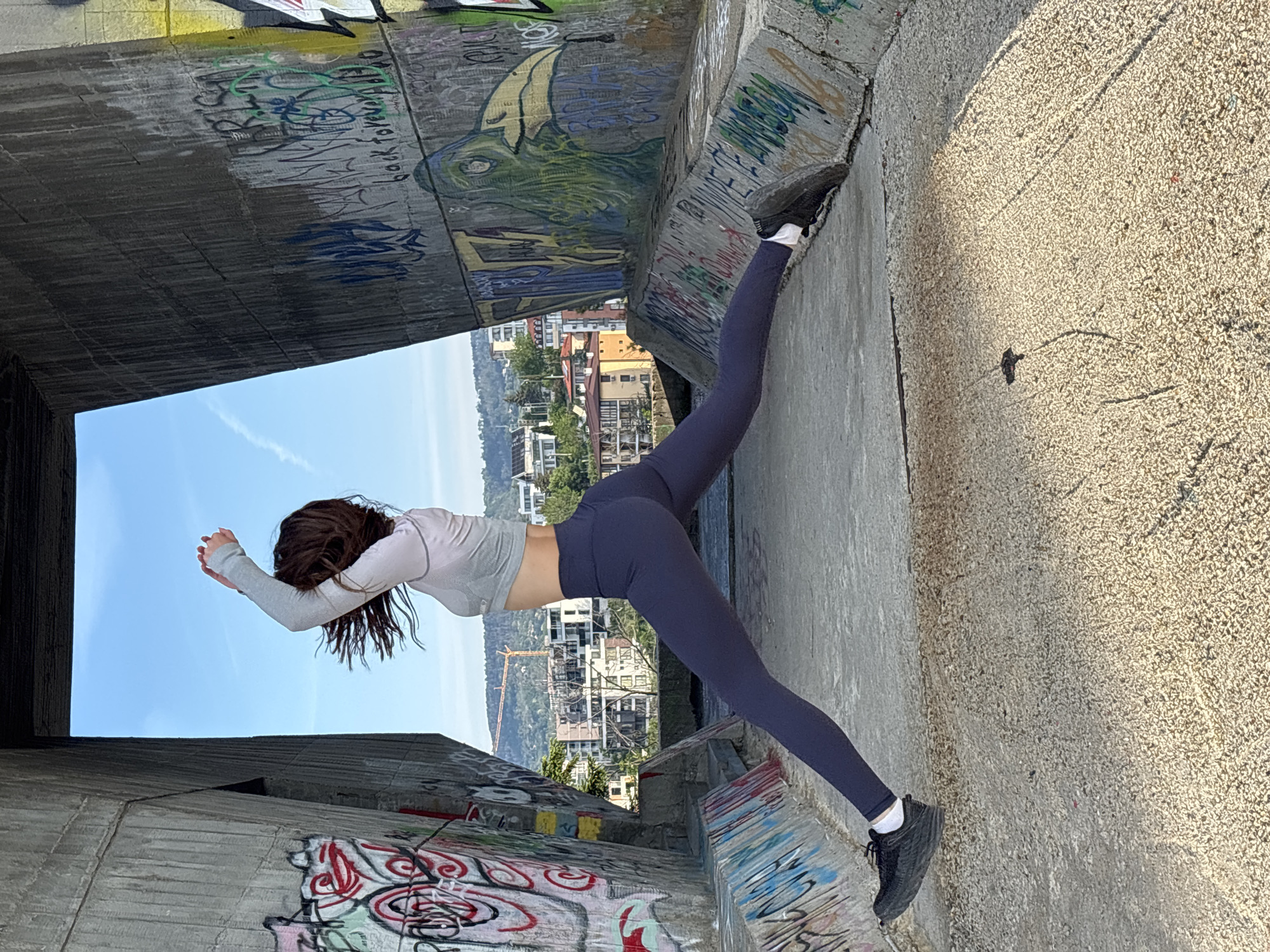 A young woman with long brown hair is performing a yoga split between two concrete bridge supports. She is wearing gray long sleeves and dark blue leggings. Graffiti covers the concrete supports. Buildings and a body of water are visible through the opening between the supports. The location is in Varna, Bulgaria, near the Asparuhov Bridge.