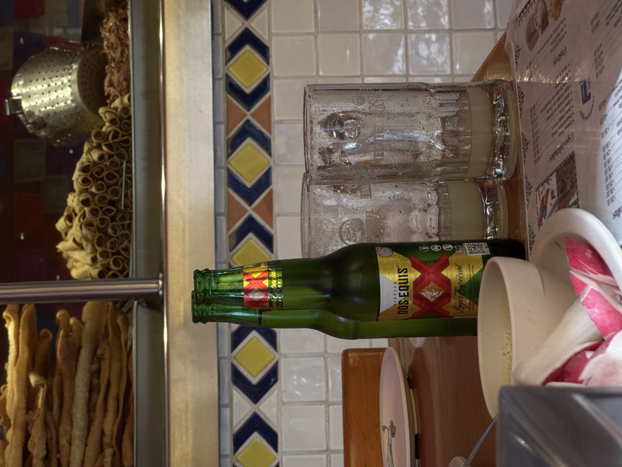 This eye-level shot, rotated sideways, features a culinary scene indoors, possibly in a restaurant in Puebla, Mexico, judging by the menu's Spanish language and cultural context. On the left side, a food display case is filled with various fried dough pastries, some cylindrical and some spiraled, likely churros or similar regional treats. The display is behind a glass partition with a metal frame and bars.
On a wooden table to the right, two condensation-covered glasses are stacked. Next to them, two green glass bottles of Dos Equis Lager Especial beer are positioned, with the brand name clearly visible. A menu with Spanish text listing dishes and prices sits partially on the table, indicating a restaurant setting. A white bowl filled with pink and white radishes is also visible, suggesting a salad or garnish preparation area.
The background features tiled walls, some with decorative diamond patterns in blue, yellow, and brown. The scene appears to be in a bustling commercial establishment, likely a restaurant or food stall in Puebla.