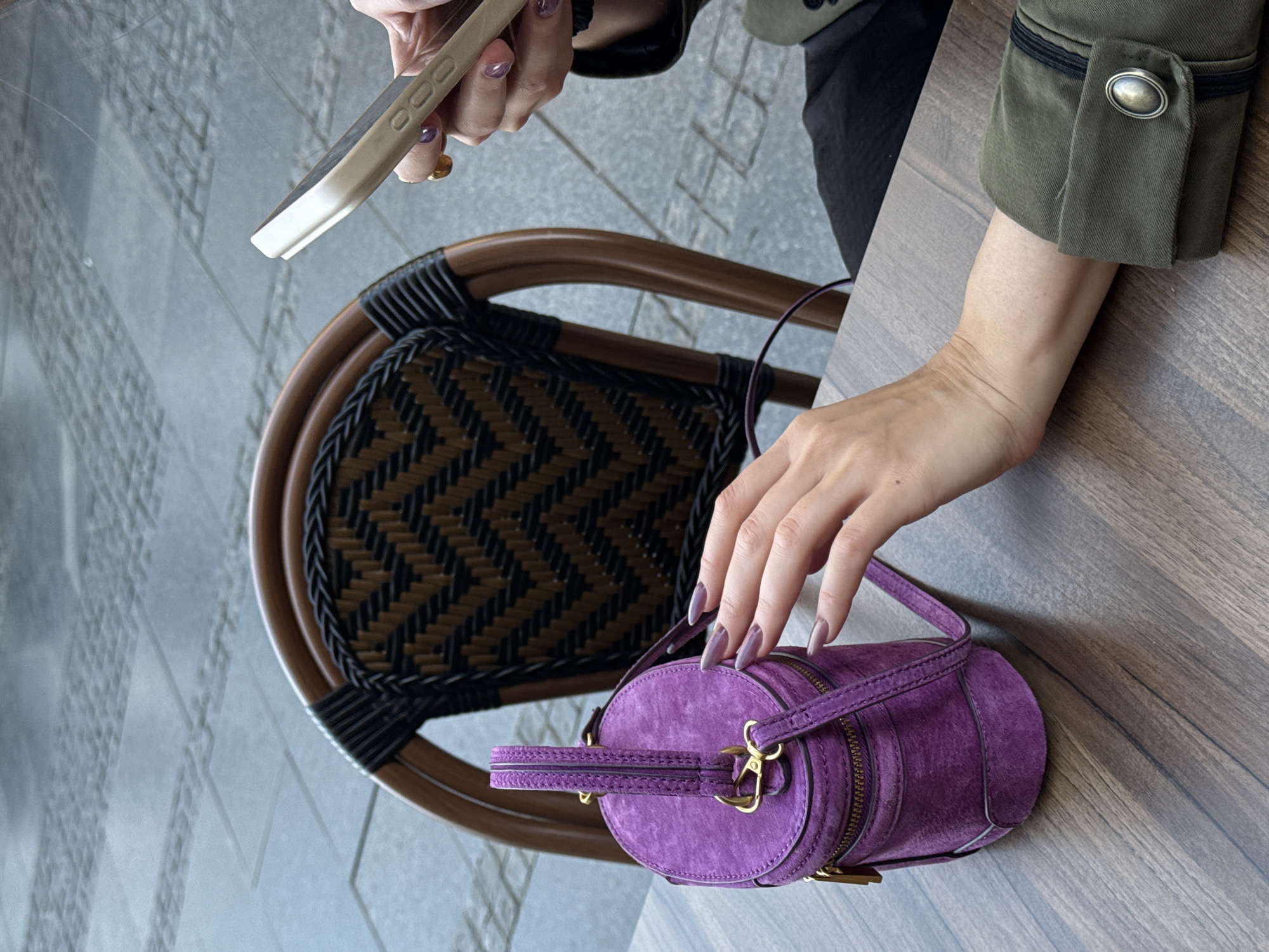 The image shows a person seated at an outdoor cafe, likely in Belgrade, Serbia, based on the presence of a wicker chair common in such settings. The person is holding a mobile phone in their left hand, while their right hand rests on a purple, cylindrical handbag placed on the wooden table. The individual is wearing a green jacket with a buttoned cuff. The cafe appears to be located on a tiled pedestrian area.