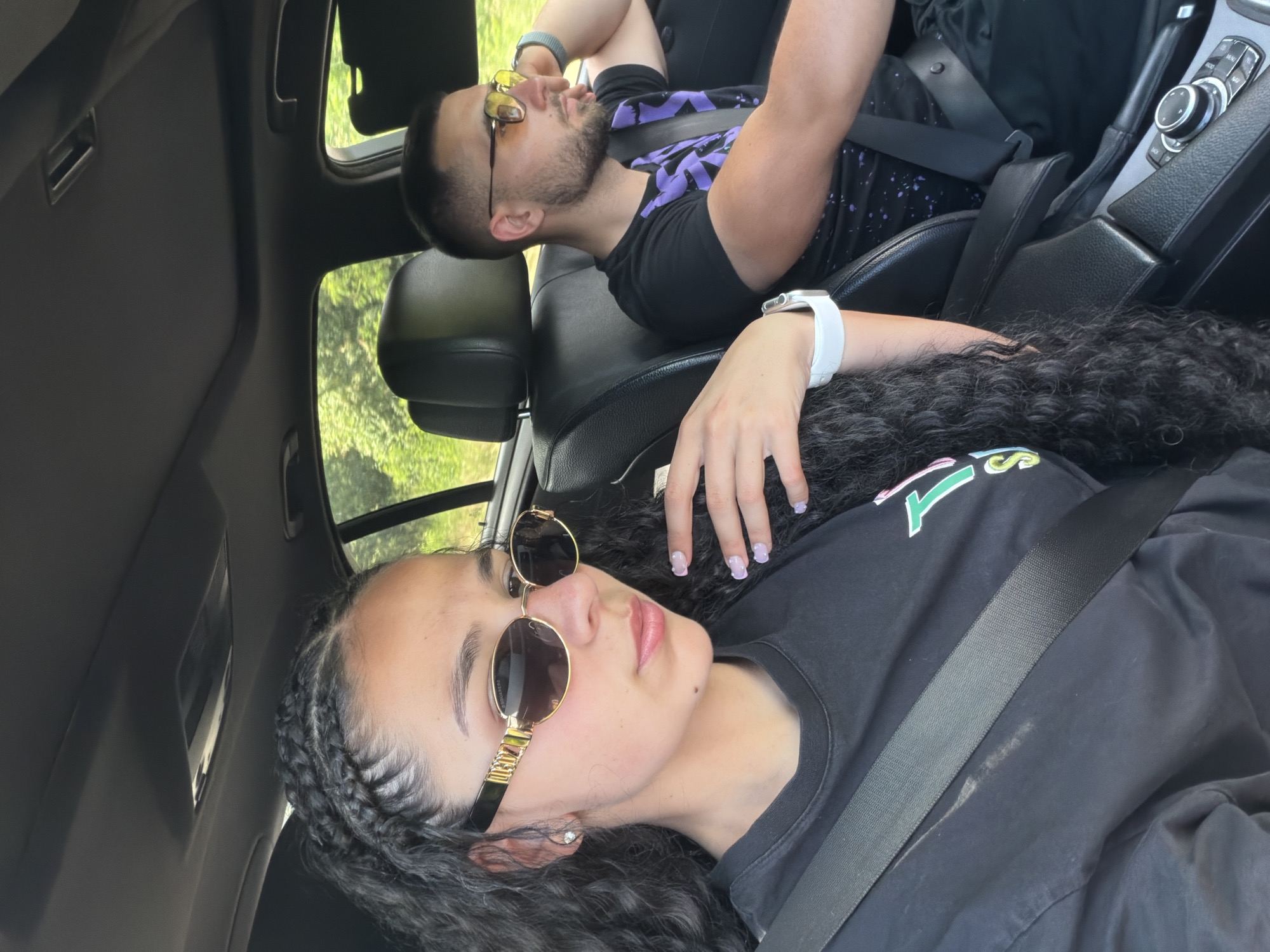 The media file shows two people in the front seats of a car. The person in the foreground is a woman with dark curly hair, wearing sunglasses and a black t-shirt. She has braids on the side of her head and a white smartwatch on her left wrist. The person in the background is a man with short dark hair and a beard, also wearing sunglasses and a black t-shirt with a purple design. He is leaning his head back, with his arms crossed. The scene appears to be during the daytime, with sunlight filtering through the car windows and trees visible outside. The overall mood is relaxed and casual. There is no visible text in the image. The location context provided is Khadzhiysko, Bulgaria.