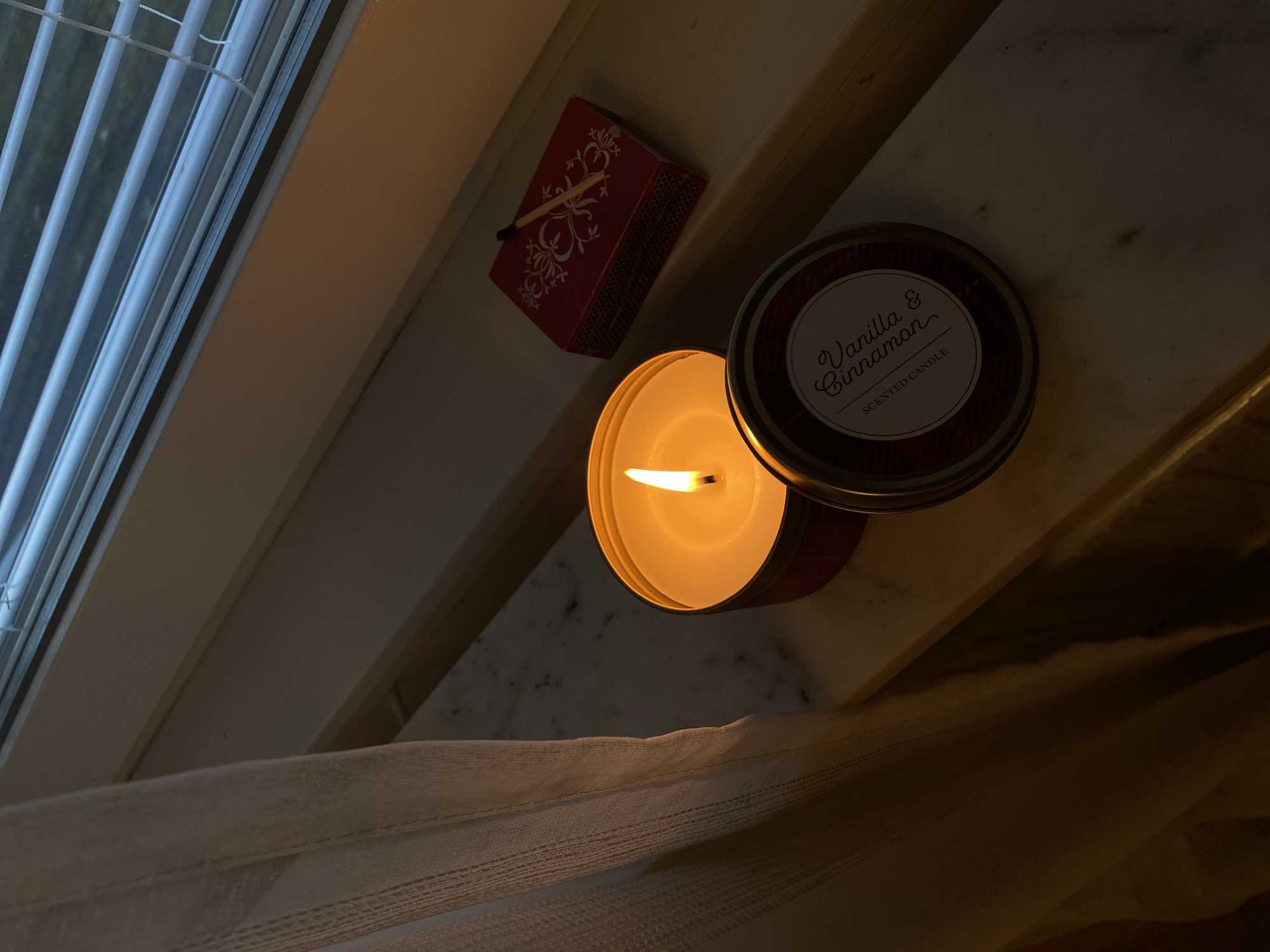 A lit "Vanilla & Cinnamon" scented candle, open with the lid beside it, sits near a matchbox with a match on top, on what appears to be a windowsill next to window blinds. A curtain is also visible. The location and city of Björklinge, Sweden are not discernible from the provided image.