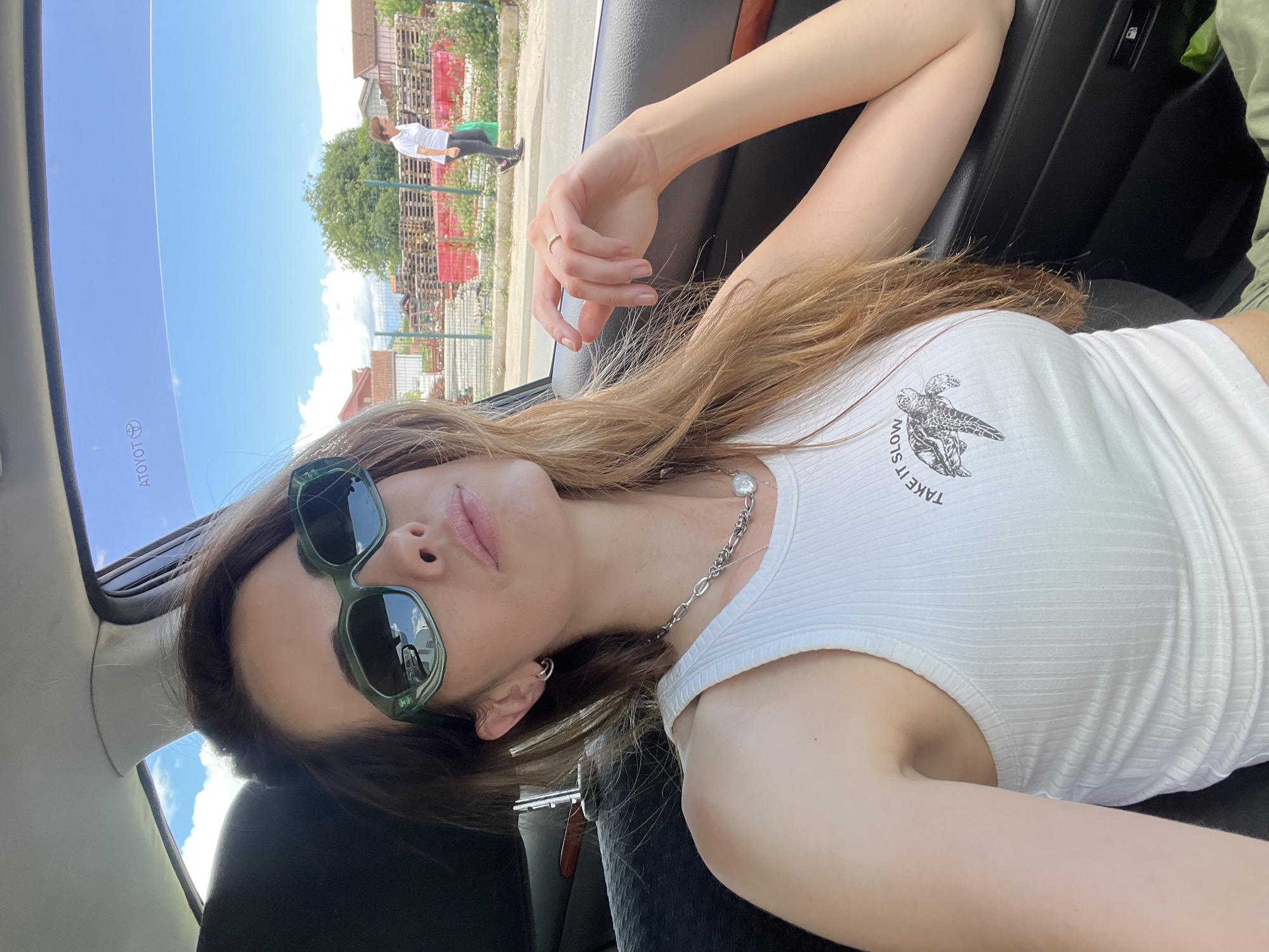 A woman reclines inside a car, taking a selfie or being photographed, with the image rotated 90 degrees counter-clockwise. She has long, light brown hair, green rectangular sunglasses, and a white ribbed crop top with a graphic of a turtle and the partially visible text "TAKE IT SLOW." A silver chain necklace with a circular pendant is visible around her neck. Her expression is relaxed, with slightly parted lips.
Through the car window, a bright, sunny outdoor scene is visible. The blue sky has a few clouds, indicating clear weather during the day. In the background, a person wearing a white shirt and dark pants stands near large stacks of wooden pallets and green fencing. Small buildings or houses and a tree are also visible in the distance. The setting appears to be an outdoor, possibly industrial or semi-rural area in Podgorica, Montenegro. Reflected on the car's interior or sunroof, the reversed and partially cropped text "TOYOTA" is discernible, suggesting the brand of the vehicle.