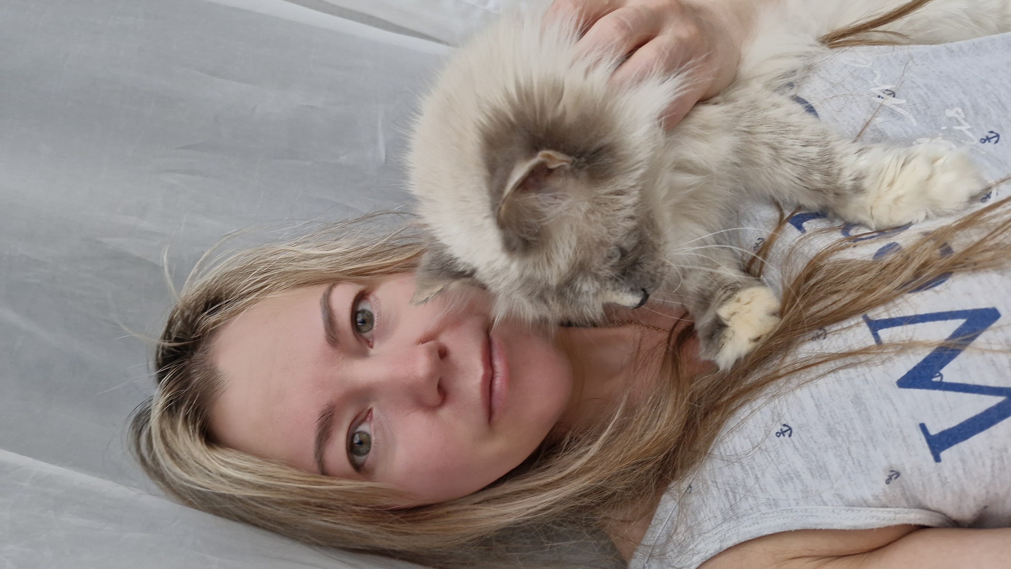 A light-skinned woman with light brown and blonde hair is lying on a white surface. A fluffy, light gray and white cat is on her chest. The woman is wearing a light gray shirt with a blue 'M' and anchor patterns on it. The image does not include any contextual clues for the city of Otchor, Russia.