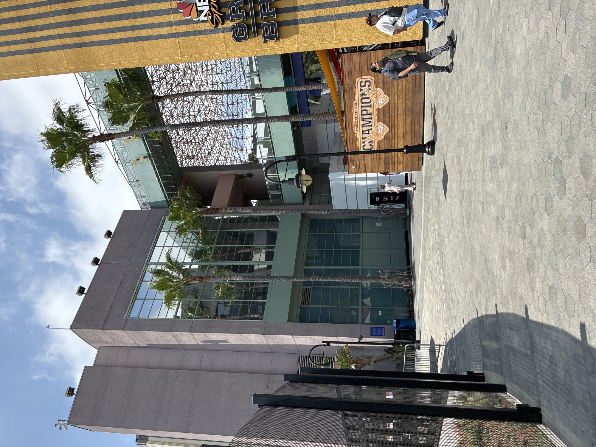 This is a medium shot, eye-level, outdoor, daytime, full-color, eye-level photograph of the Universal Studios Hollywood theme park in Universal City, California. The scene depicts an outdoor plaza area. To the left is a tall, gray building with a large, multi-paned glass facade reflecting palm trees. In front of the building is a black metal fence. Several palm trees are visible throughout the image. To the right of the gray building is a tall, yellow building with vertical dark gray stripes. A sign on the yellow building reads "NBC SPORTS GRILL" with a logo above it. Below the NBC sign, a horizontal wooden sign reads "#NBCGRILLBREW CHAMPIONS". Two men are walking past the wooden sign from right to left. One man is wearing a grey t-shirt and grey pants, and the other is wearing a white t-shirt and blue pants. A woman in a light-colored outfit is walking away from the camera in the background to the left of the wooden sign. The ground is paved with hexagonal grey stones. In the upper portion of the image, a covered walkway or bridge is visible between the gray and yellow buildings, supported by a lattice-like structure. The sky is blue with scattered white clouds.