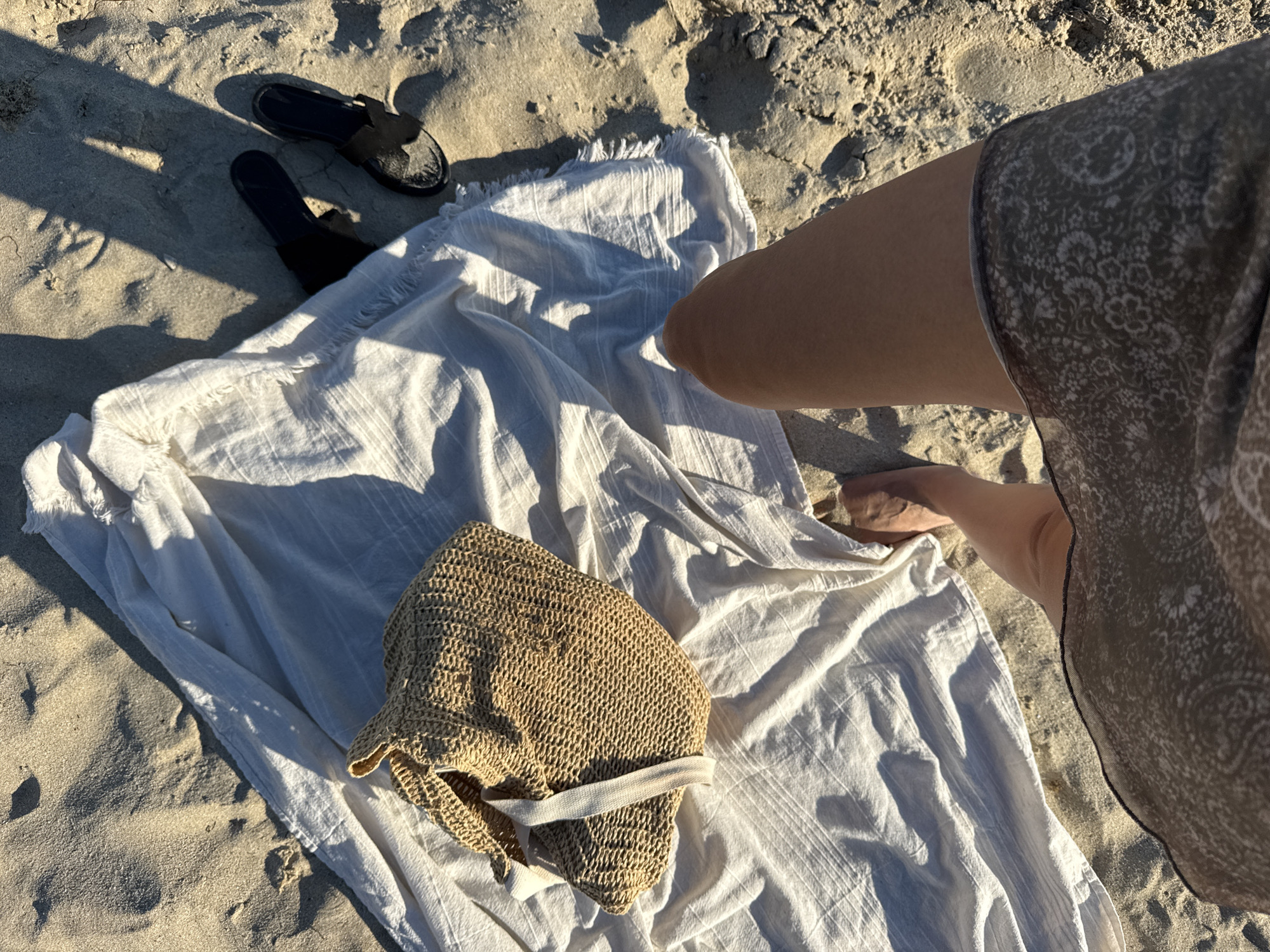 This is a high-angle, medium shot of a person's legs and a beach blanket on a sandy beach in Nesebar, Bulgaria. The person is wearing a patterned skirt and is standing barefoot. To the left of the person is a white textured blanket with fringe detail. On the blanket is a woven straw bag with white straps. To the right of the blanket are a pair of black slide sandals. The sand is light brown with visible footprints and shadows cast by an unseen object. The sun is shining brightly.