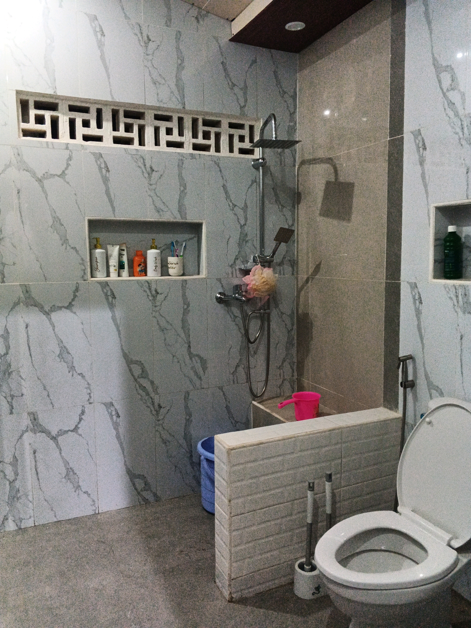 This is a forensic, matter-of-fact description of a bathroom scene in Sirau, Indonesia. The bathroom features marble-patterned tiles on the walls and a gray, textured floor. On the left wall, there is a recessed shelf containing several bottles of toiletries, including shampoo and body wash, as well as a toothbrush holder with toothbrushes. Above this shelf is a decorative rectangular vent with a geometric pattern.
In the center of the room, a shower head with a hose and a pink loofah is visible. Next to the shower, a small pink bucket sits on a ledge. A blue plastic bucket is also visible behind the ledge. To the right of the shower area, a partition wall with a textured white brick pattern separates the shower from the toilet area. A bidet sprayer is mounted on the wall next to the partition.
The right side of the image shows a white toilet with the lid and seat up. Next to the toilet, a toilet brush and holder are positioned on the floor. On the far right wall, there is another recessed shelf containing a green bottle, likely cleaning fluid or soap. The lighting in the bathroom appears to be artificial, with a ceiling light visible in the upper right corner. There are no people visible in the image.