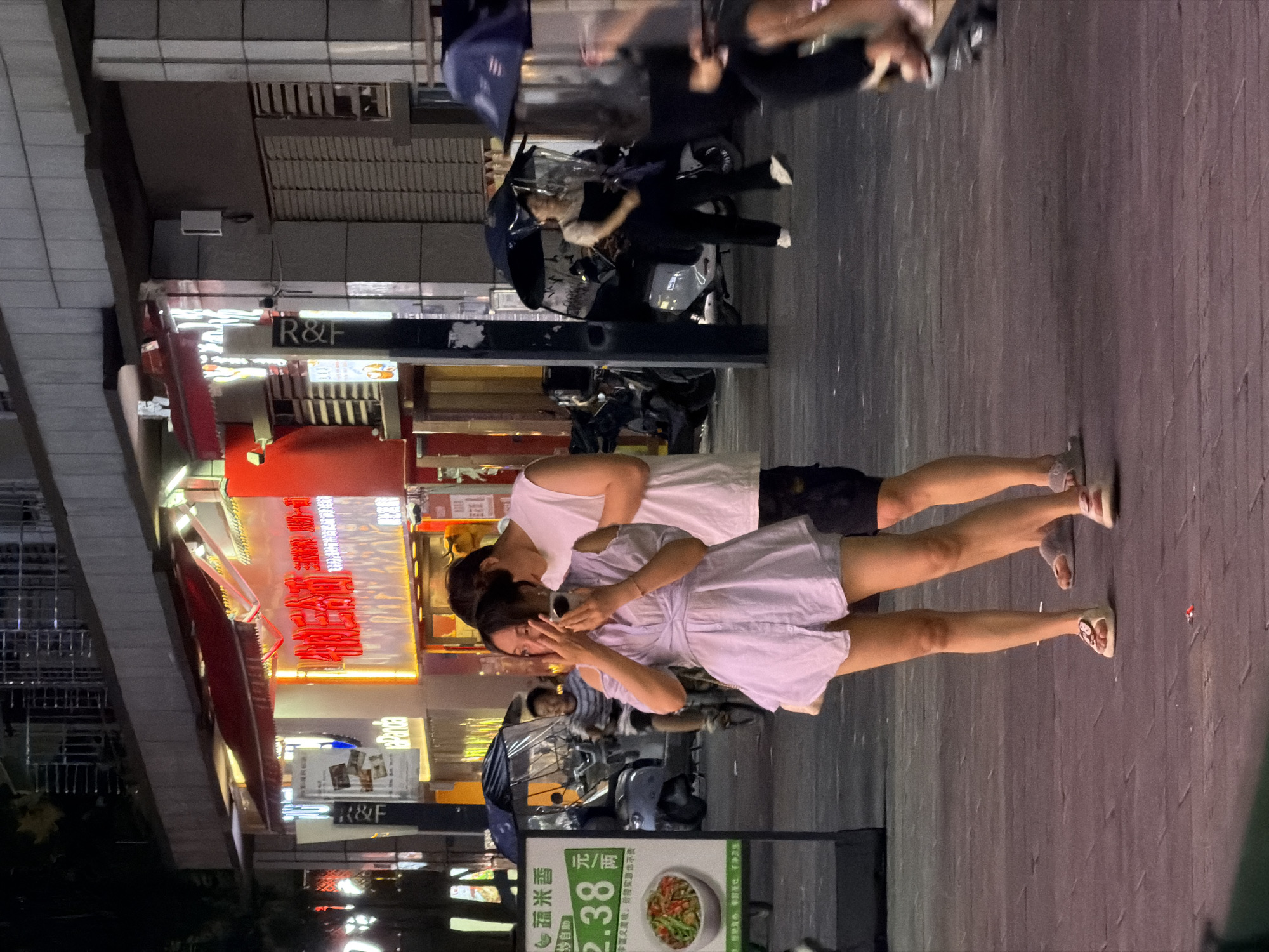The image, taken at an evening, features a street scene, likely in the city of Chongqing High-tech Zone, China. The scene is well-lit by streetlights and illuminated shop signs.
A couple, likely of Asian descent, is in the foreground. The woman wears a light-colored dress and the man a white top and shorts. They are close to each other, possibly looking at a phone. They wear sandals.
Behind the couple, there is a street scene with parked motorbikes. A sign with Chinese characters advertises a restaurant or food stall. The sign also contains prices. A variety of shop signs with both Chinese and English characters line the street. The architectural style of the buildings suggests a modern urban environment.
