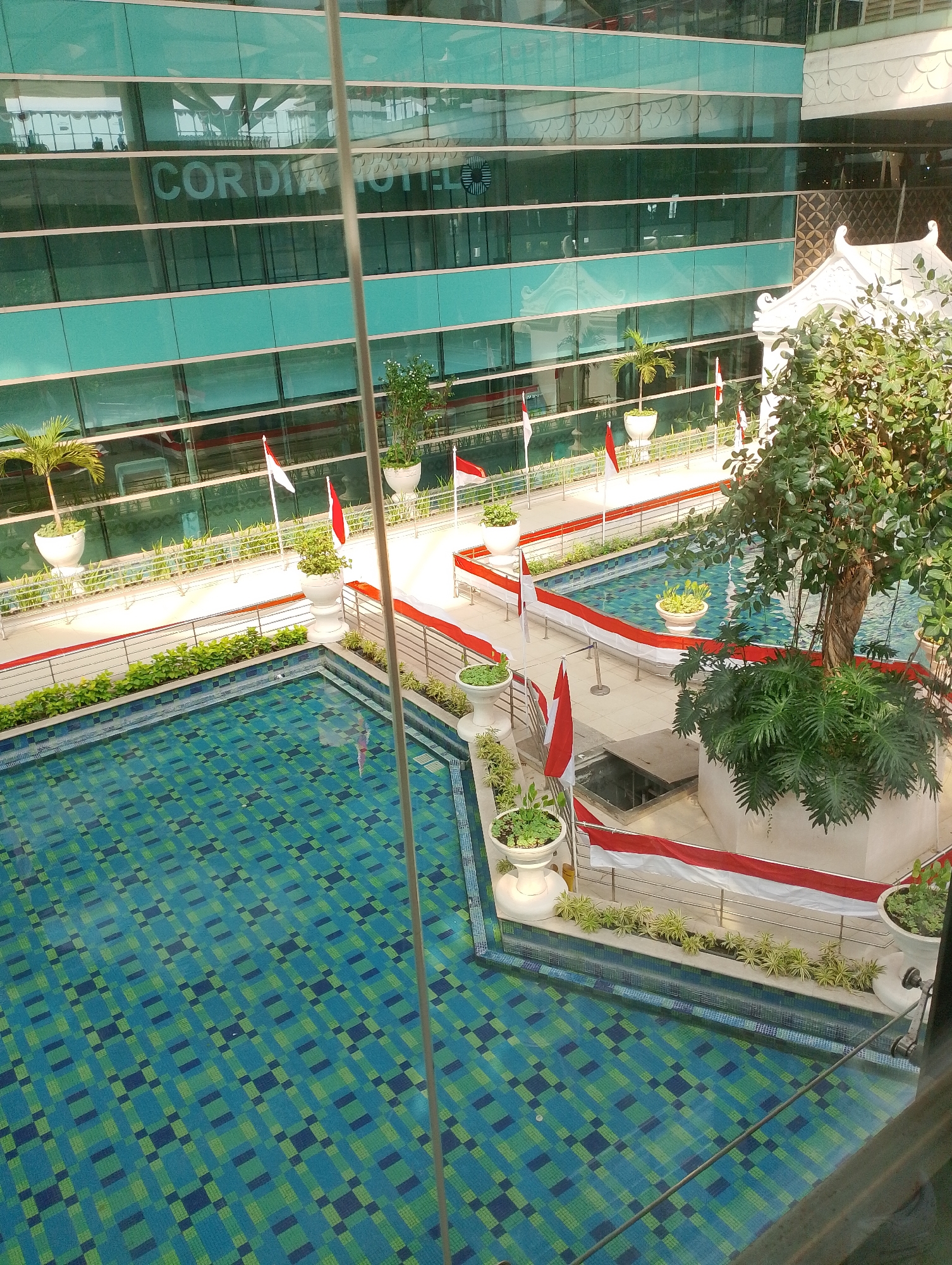 This image captures an outdoor swimming pool area, likely a hotel or resort, with a distinctive Indonesian theme, suggested by the red and white flags decorating the perimeter. The pool itself is rectangular with a patterned mosaic tile bottom in shades of blue and green. Surrounding the pool is a tiled deck with planters containing greenery. In the background, a modern building with a glass facade dominates the scene, reflecting the sky and surrounding structures. The reflection on the building's glass indicates "CORDIA HOTEL" and other corporate signage. The overall impression is a blend of modern architecture and a tropical outdoor amenity, with a patriotic decorative touch.