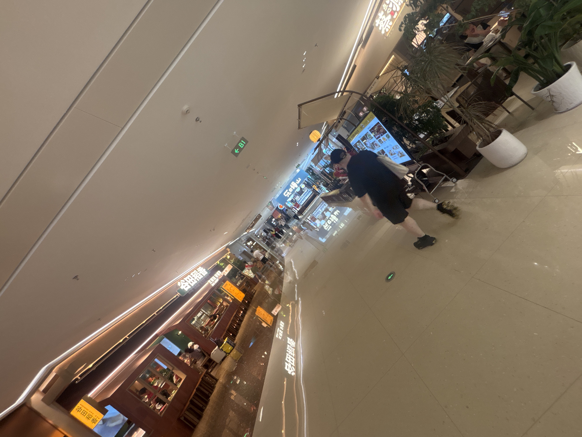 The image shows an interior view of a shopping mall hallway in Haizhu District, China. A person in a black t-shirt and shorts is walking away from the camera, with their back to the viewer. The hallway is well-lit with overhead fluorescent lighting and decorative LED strip lighting along the ceiling and walls. Several storefronts are visible, including restaurants with illuminated signage displaying Chinese characters. Some storefronts have seating areas with customers. A digital menu board is positioned near the center of the image. Potted plants are placed along the walkway. The floor is polished and reflective, showing reflections of the ceiling lights and storefronts. A directional sign indicating "B1" with an arrow is mounted on the ceiling. The perspective is tilted, giving a dynamic feel to the scene.