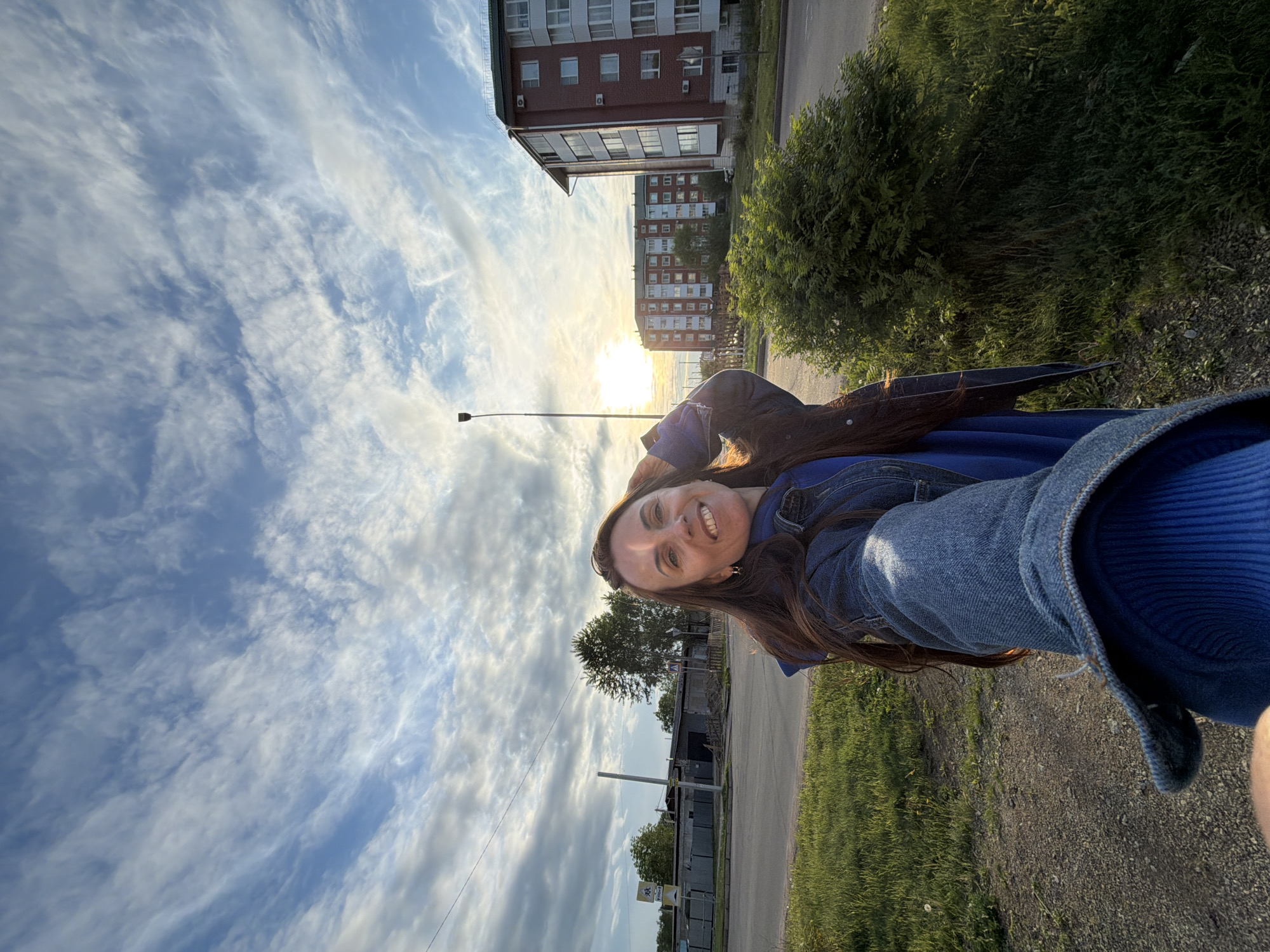 This image depicts an outdoor scene in Birobidzhan, Russia, captured during the late afternoon or early evening, characterized by the low sun on the right side of the frame. An adult female is positioned in the foreground, smiling directly at the camera, holding the device with her extended right arm in a selfie orientation. She has long, dark brown hair, fair skin, and is wearing a blue long-sleeved top beneath a blue denim jacket. Her left hand is raised to her head, near her hair. Strong backlighting from the sun illuminates her hair and the right side of her body.
The ground in the immediate foreground consists of a dirt and gravel path, transitioning into green grass with sparse vegetation. An asphalt road spans the midground horizontally, flanked by grassy verges. On the right side of the road, a dense, green bush is present. To the left of the woman, beyond the road, there are several low-rise industrial-style buildings with grey facades and fencing, along with mature green trees. A yellow sign with text on a blue background is visible near these structures. To the right of the woman and the road, a tall, black street light pole with a single luminaire stands. Further right, a multi-story residential building constructed of red brick with white window frames and balconies is prominent. Another similar red-brick building is visible in the background behind it, partially obscured by the bright sun. The sky is predominantly blue with scattered white, wispy cirrus and cumulus clouds.