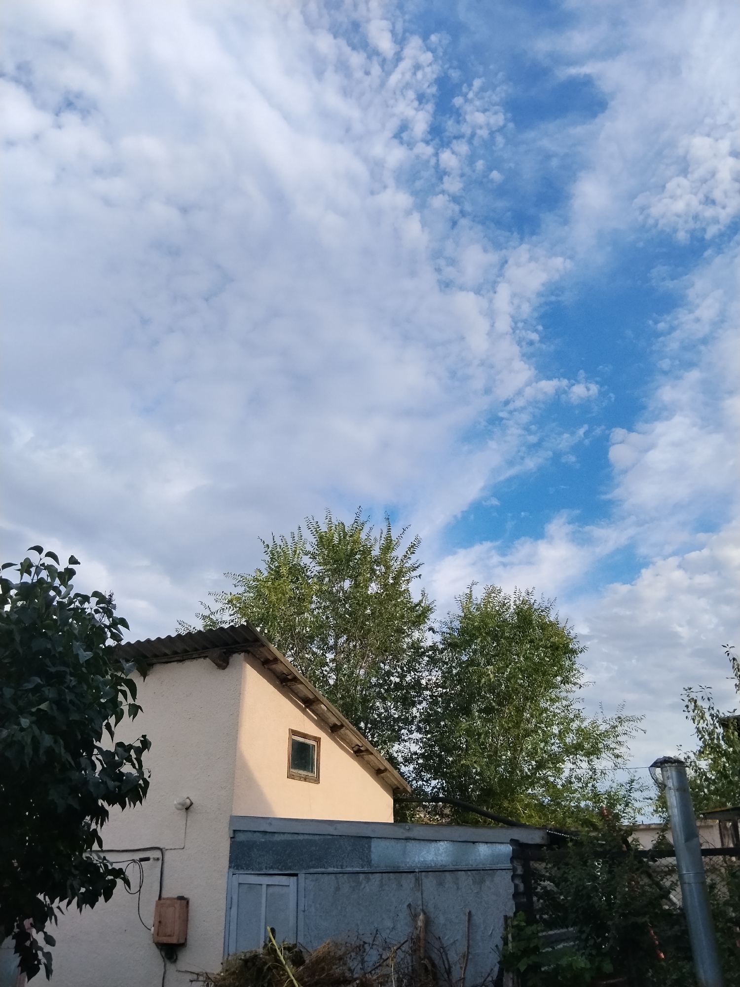 The image depicts a suburban scene in Kokshetau, Kazakhstan, under a sky filled with scattered white clouds against a bright blue background. In the foreground, a two-story structure with a corrugated metal roof is visible. The upper part of the building is cream-colored, featuring a small, square window with a dark frame. Below this, a metal garage door is present, with a lighter blue door visible behind it. To the left of the building, dark green foliage from a tree partially obscures the scene. To the right and behind the building, several tall, leafy green trees are seen, with a metal chimney pipe standing tall on the right edge of the frame. The overall impression is one of a rural or semi-rural residential area. No people are visible in the image.