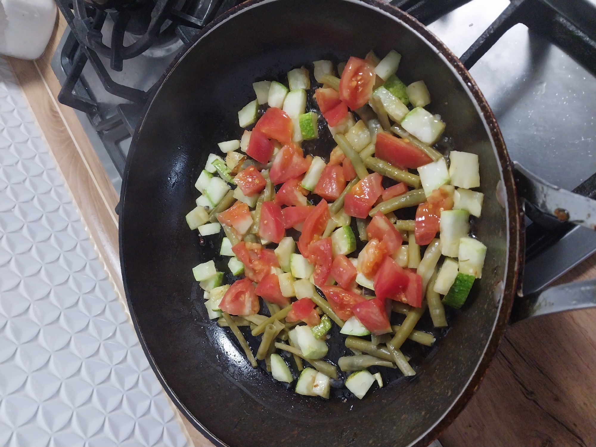 In an indoor kitchen setting, a black frying pan is placed on a gas stove. The pan contains chopped vegetables, primarily zucchini and tomatoes, along with green beans. The vegetables appear to be in the process of being cooked, with some moisture visible in the pan. The stovetop and surrounding countertop are visible, along with a backsplash with a textured pattern. The image does not provide any information that would allow for identification of the city of Dnipro, Ukraine.