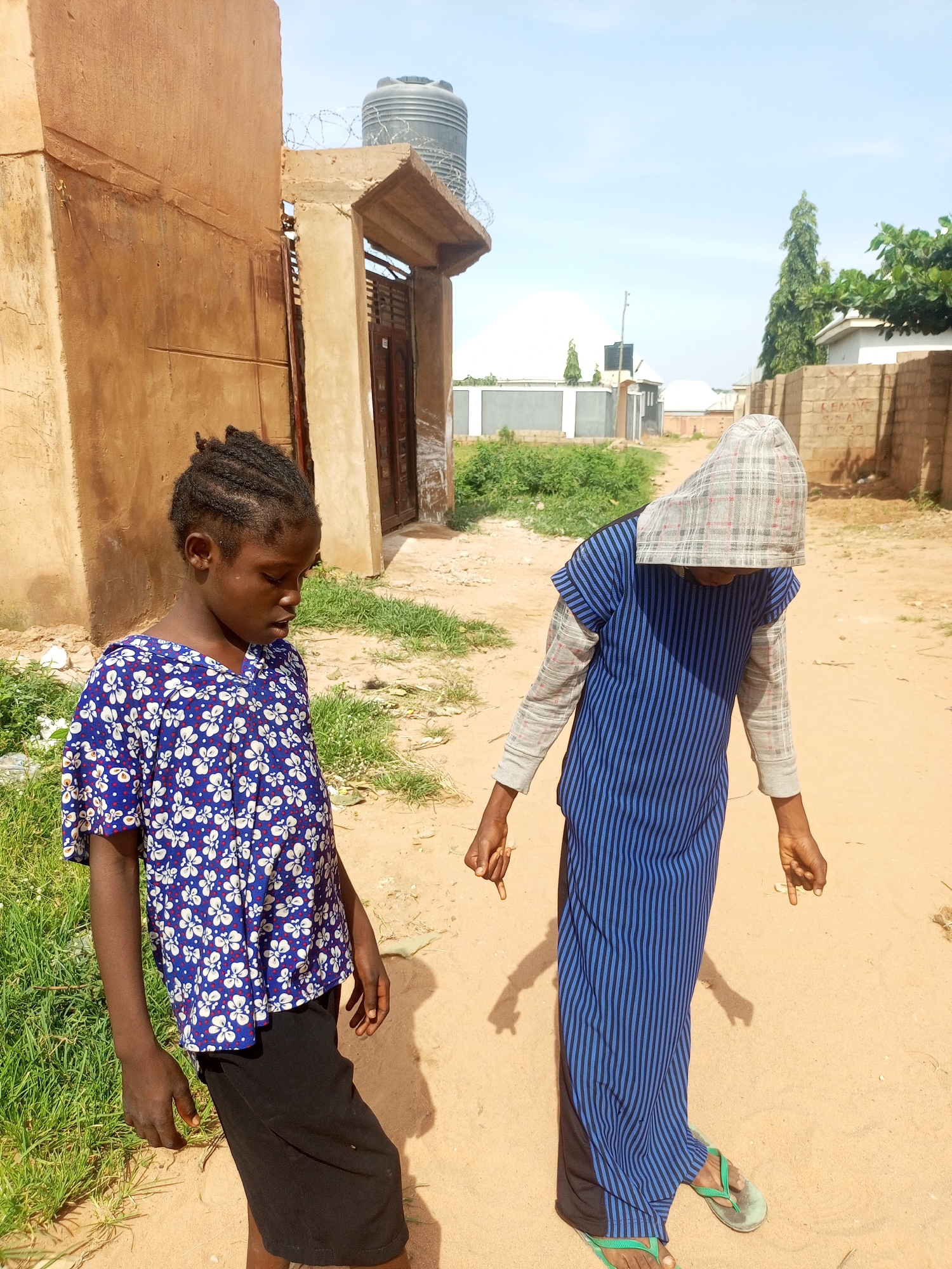 In Tempure, Nigeria, on a sunny day, two young girls are standing in a dusty, unpaved area with sparse green grass. The girl on the left, facing forward with her head tilted down, wears a blue short-sleeved shirt with a white and red floral pattern and black shorts. Her braided dark hair is pulled back. The girl on the right, facing away from the camera with her head covered by a checkered cloth, wears a long blue and black striped dress over a gray long-sleeved shirt and dark pants. She is also wearing light green flip-flops. Behind them, a section of a light brown building with a dark wooden door and a water tank on top is visible, along with a fence and a few trees in the distance. The ground is sandy and dry, with some debris scattered around.