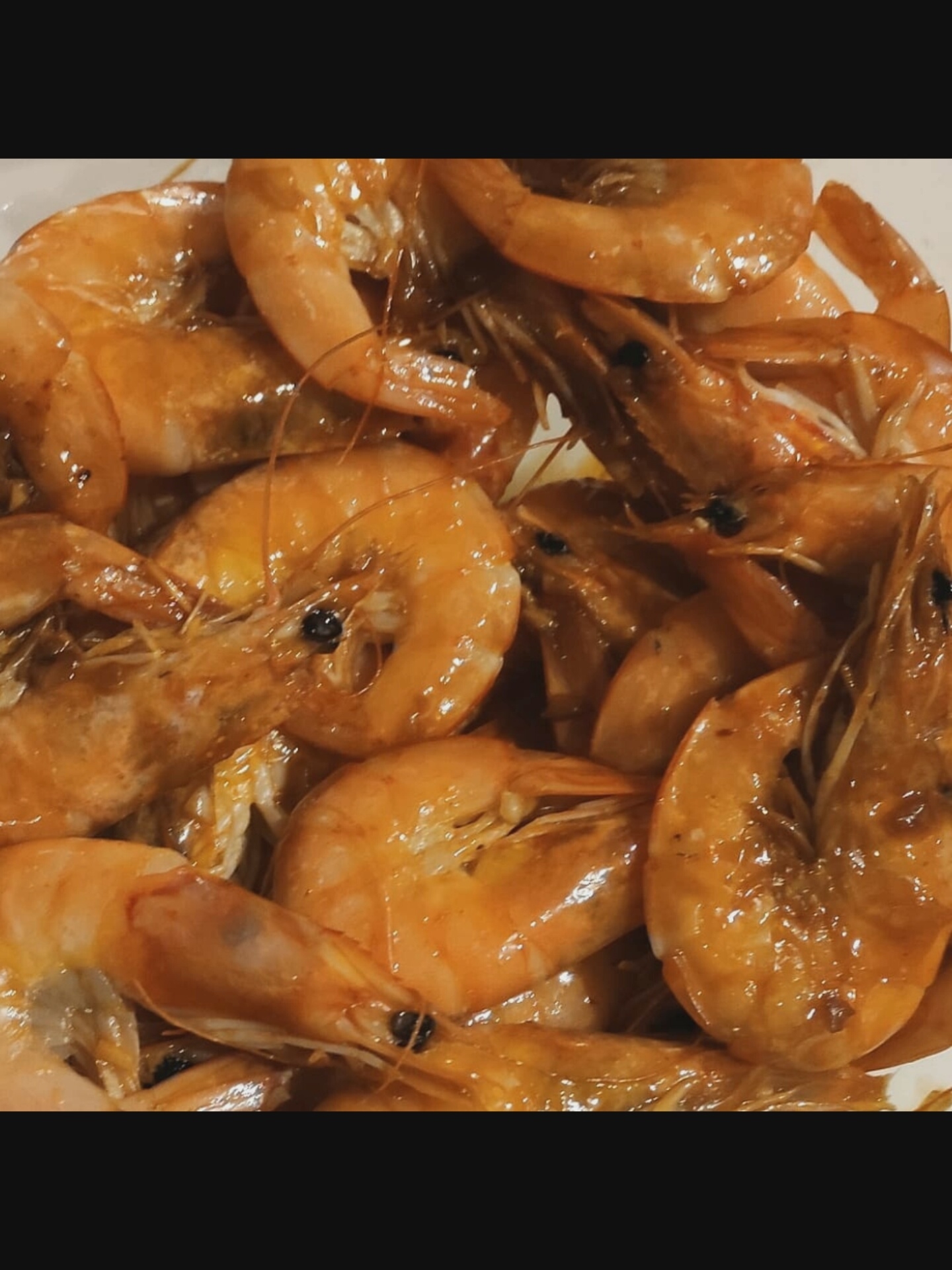A close-up, overhead view of a bowl filled with cooked shrimp. The shrimp are a bright orange-pink color, and appear to be glazed with a sauce. They are piled high in the bowl, with some overlapping each other. The background is out of focus, but appears to be a light-colored surface. There are no people or animals visible in the image. The location in the city of Saint-Août, France is not determinable from the image.