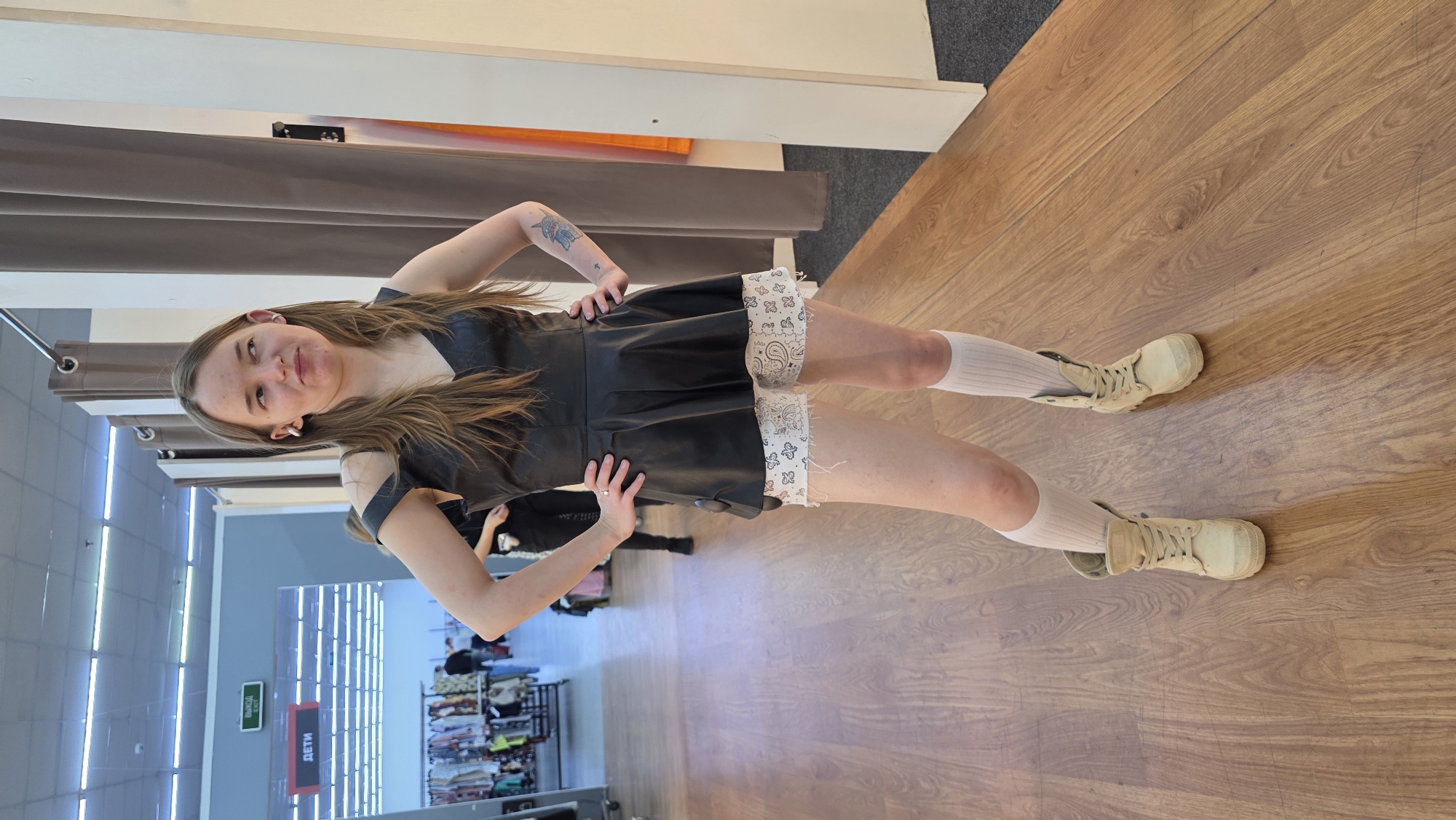 A young woman is standing indoors, posing for a photograph, likely in a clothing store or a fitting room area. She is smiling and looking towards the camera, with her hands on her hips. Her long, light brown hair is falling around her shoulders. She is wearing a sleeveless black dress, possibly made of faux leather, which features a distinctive white trim with a bandana-like pattern along the hem. Underneath, she wears white socks, possibly knee-high or calf-high, paired with light beige, lace-up combat boots. A tattoo is visible on her left arm.
The setting features a light brown wooden floor and grey-brown fitting room curtains behind her. In the background, to the left, there are clothing racks with various garments, suggesting a retail environment. The lighting is artificial, illuminating the space with overhead strip lights visible on the ceiling.
Visible text on signs in the background confirms the location as a store. A red sign with white text reads "ДЕТИ" (DETI), which translates to "CHILDREN." Another green sign with white text says "Выход" (Vykhod), meaning "Exit." The overall impression is a casual, cheerful moment captured during a shopping trip.