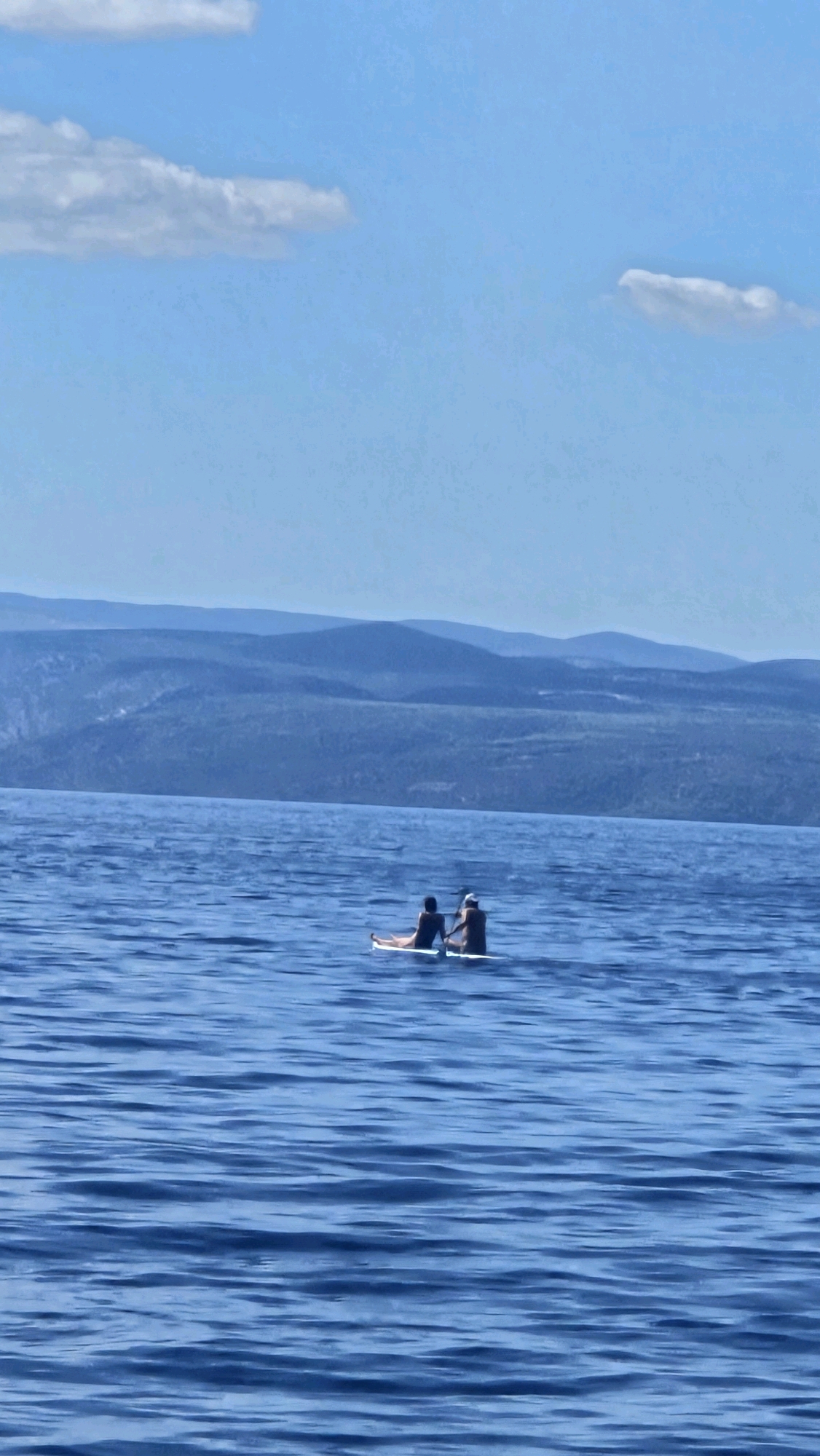 The image depicts two individuals on paddleboards in the Adriatic Sea, with the coastline of Općina Podgora, Croatia, visible in the background. The water is a deep blue with gentle ripples. In the foreground, two people are seated on separate paddleboards. The person on the left is facing away from the camera, with their legs extended. The person on the right is also facing away, holding a paddle. They appear to be in a relaxed pose. The middle ground features a vast expanse of blue water, meeting a hazy, layered landscape of mountains and hills that form the coastline. The sky above is a clear, pale blue, with a few scattered white clouds. The overall scene suggests a peaceful and scenic day on the water in a coastal Croatian location.