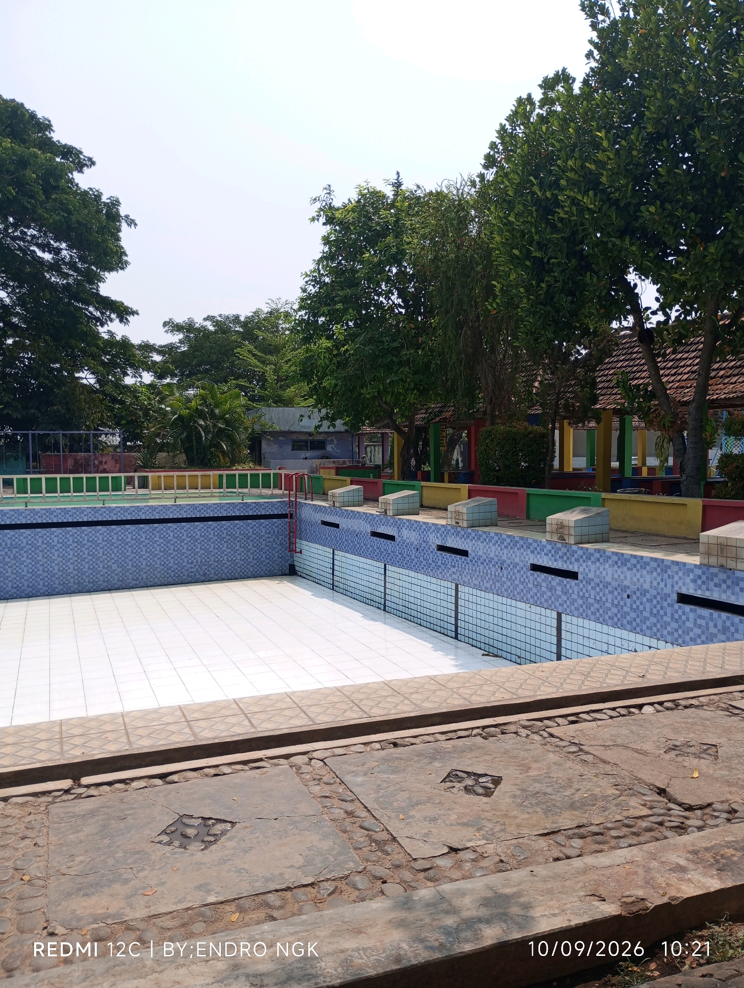 An empty, tiled swimming pool is the central focus of this image. The pool has a white tiled bottom and light blue tiled walls. A red ladder is visible on the right side of the pool. The pool area is surrounded by a decorative border and a fence. In the background, there are trees, buildings with red tile roofs, and a bright, clear sky. The foreground shows a cracked concrete pavement with stones embedded in it. The image is dated 10/09/2026 at 10:21 and is attributed to REDMI 12C | BY:ENDRO NGK. The specific location is stated to be the city of Sombron, Indonesia.