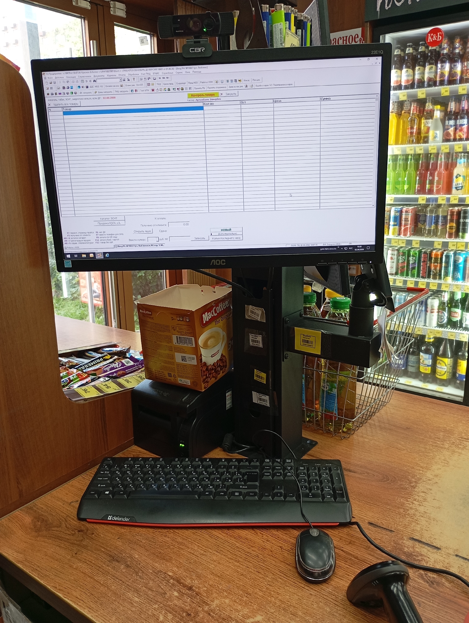 This is a high-angle, medium shot of a retail checkout counter in Moscow, Russia. The scene is dominated by a computer monitor displaying a point-of-sale (POS) software interface. To the left of the monitor is a black and red keyboard, and to the right is a black computer mouse. In front of the monitor, on the counter, is a cash register and a box of "MacCoffee." Behind the monitor, a webcam is mounted on top of a shelf, and various lighters are visible on the shelf. A refrigerator filled with drinks is positioned in the background to the right of the counter. The counter surface is made of wood. The image appears to have been taken during daylight hours.