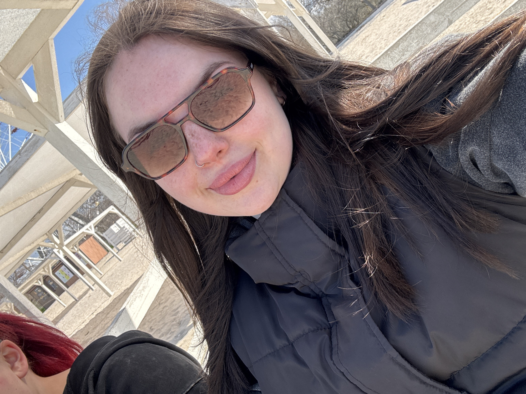 The image shows a woman with long, brown hair and sunglasses taking a selfie. She has a nose ring and is wearing a black puffer jacket over a gray top. In the background, there's a white, geometric structure, likely part of a building or pavilion, and beyond that, a glimpse of what appears to be beach scenery. At the bottom left corner of the image, a small portion of another person with red hair is visible.
Based on the context provided (Chornomorskoe, Ukraine), the photo was likely taken near the beach or seaside promenade in that city. The white structure in the background may be part of the local architecture or infrastructure near the coast.