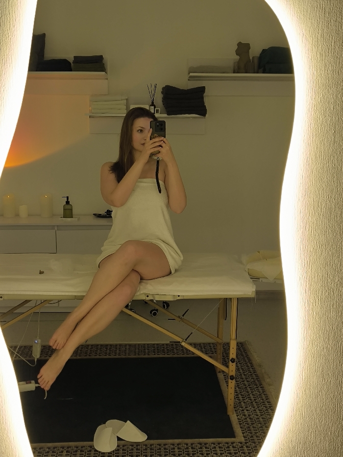 The image depicts an indoor room, identified as a spa or massage therapy setting in Vinnytsia, Ukraine. A female subject with brown hair, wrapped in a white towel, is seated on a cream-colored massage table. Her bare legs are crossed at the knee, extending leftward towards the lower left corner of the frame. She holds a dark-colored smartphone with both hands, capturing her reflection in a large, irregularly shaped mirror with an illuminated right edge. In the background, white walls feature multiple floating shelves displaying neatly folded dark and light-colored towels. A white cabinet or counter surface to the left holds two white pillar candles and a green pump bottle. To the right, a reed diffuser and a light brown decorative object are present on a shelf. The floor beneath the massage table is covered by a patterned rug, and two white disposable slippers are positioned on the rug near the subject's feet. The room is softly lit with warm, ambient lighting.