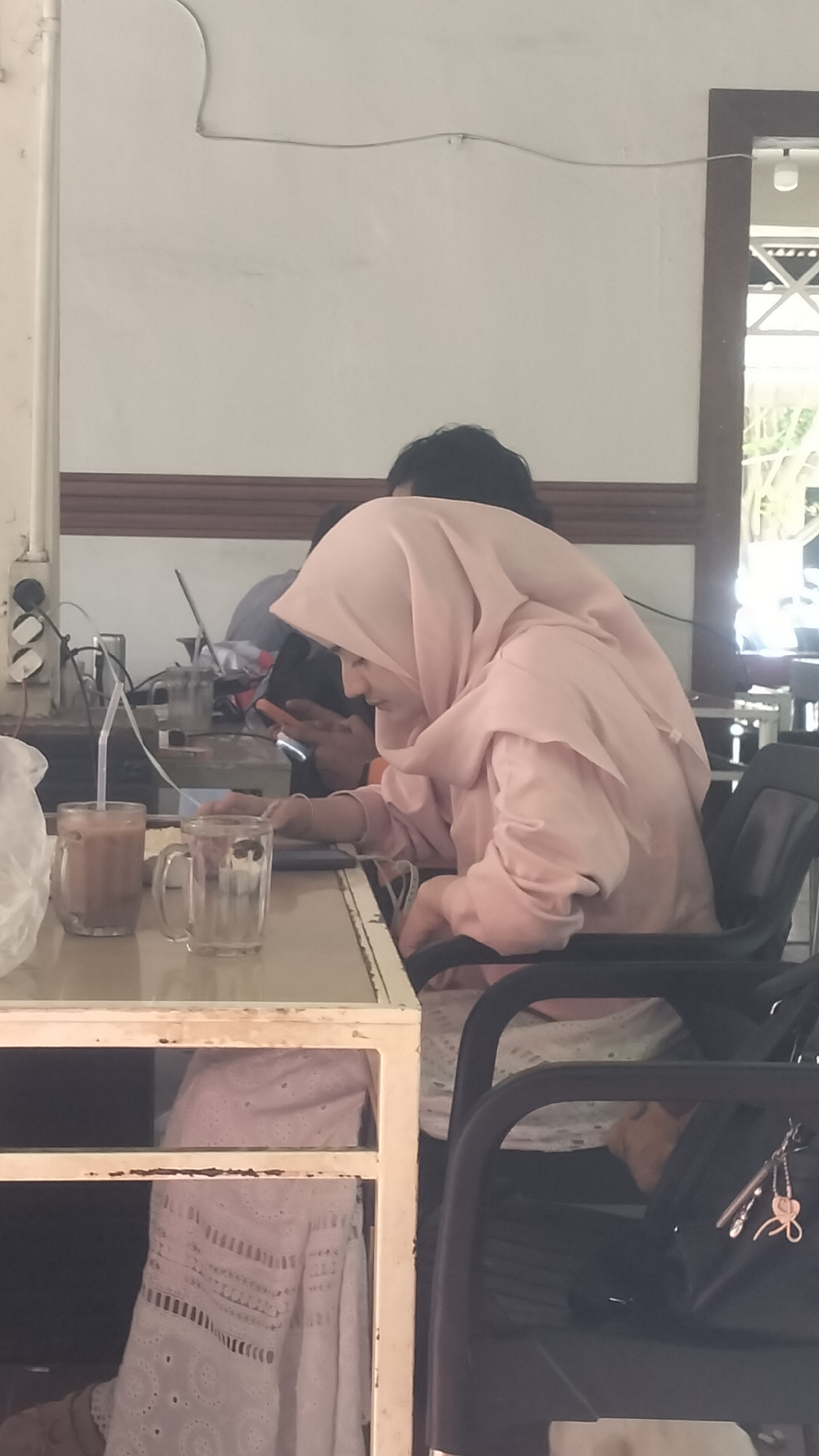 A medium shot captures a profile of a young woman wearing a light pink hijab and a matching outfit. She is seated at a table, her head bent down as she looks at her phone. Beside her is a clear glass with a straw and a liquid, and an empty clear glass. Another person, also wearing a light pink hijab, is partially visible in the background, also looking at their phone. The scene appears to be a cafe or a restaurant, indicated by the presence of drinks and tables. The background shows a white wall with some wires and electrical outlets. A window is visible on the right side, offering a glimpse of the outdoors. The overall atmosphere is casual and relaxed, with the individuals engrossed in their devices. The location is identified as Lhokseumawe, Indonesia.