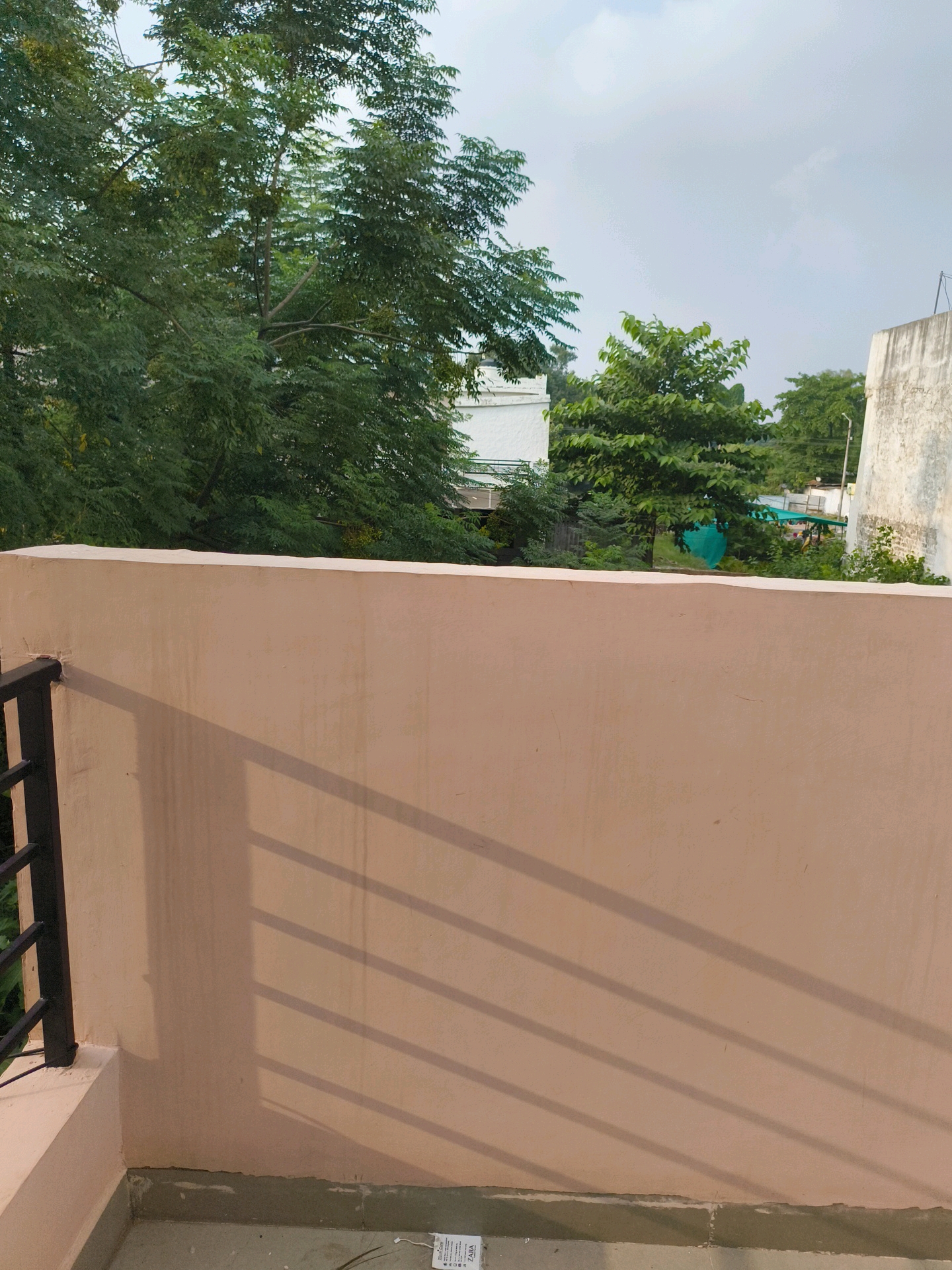 The image shows a balcony in Mohali, India, with a light pink wall dominating the foreground. Diagonal shadows cast by a railing are visible on the wall. Beyond the railing, lush green trees and some buildings are visible under a cloudy sky. The balcony floor is tiled. There are no people or animals present in the image.