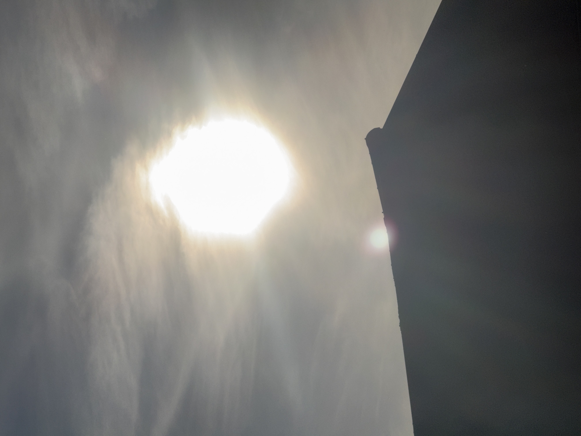 The image captures a view of the sky with the sun prominently featured, partially obscured by thin clouds. The sun appears as a bright, hexagonal shape with noticeable rays extending outwards, creating a halo effect. Below the sun, the silhouette of a dark, sloping rooftop is visible against the sky. A small, bright lens flare is also present near the horizon, to the left of the center. The overall lighting suggests either sunrise or sunset. The image was taken in Bad Salzschlirf, Germany, specifically depicting the sky above a residential building. No people are visible in the scene.