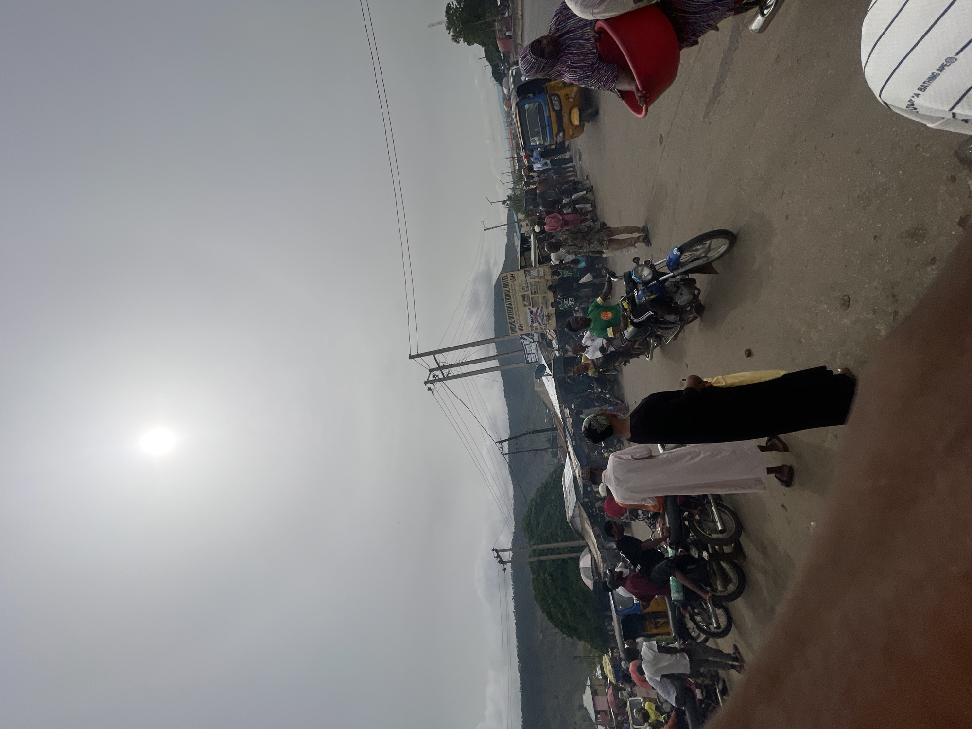 The image depicts a busy street scene in Lokoja, Nigeria. A mix of pedestrians, motorcyclists, and a yellow "keke" (auto rickshaw) occupy the road. Several people are dressed in traditional Muslim attire. The environment features utility poles and wires, and a sign for "Omide International Hotel" is visible. In the background, a hillside can be seen. The sky is overcast.