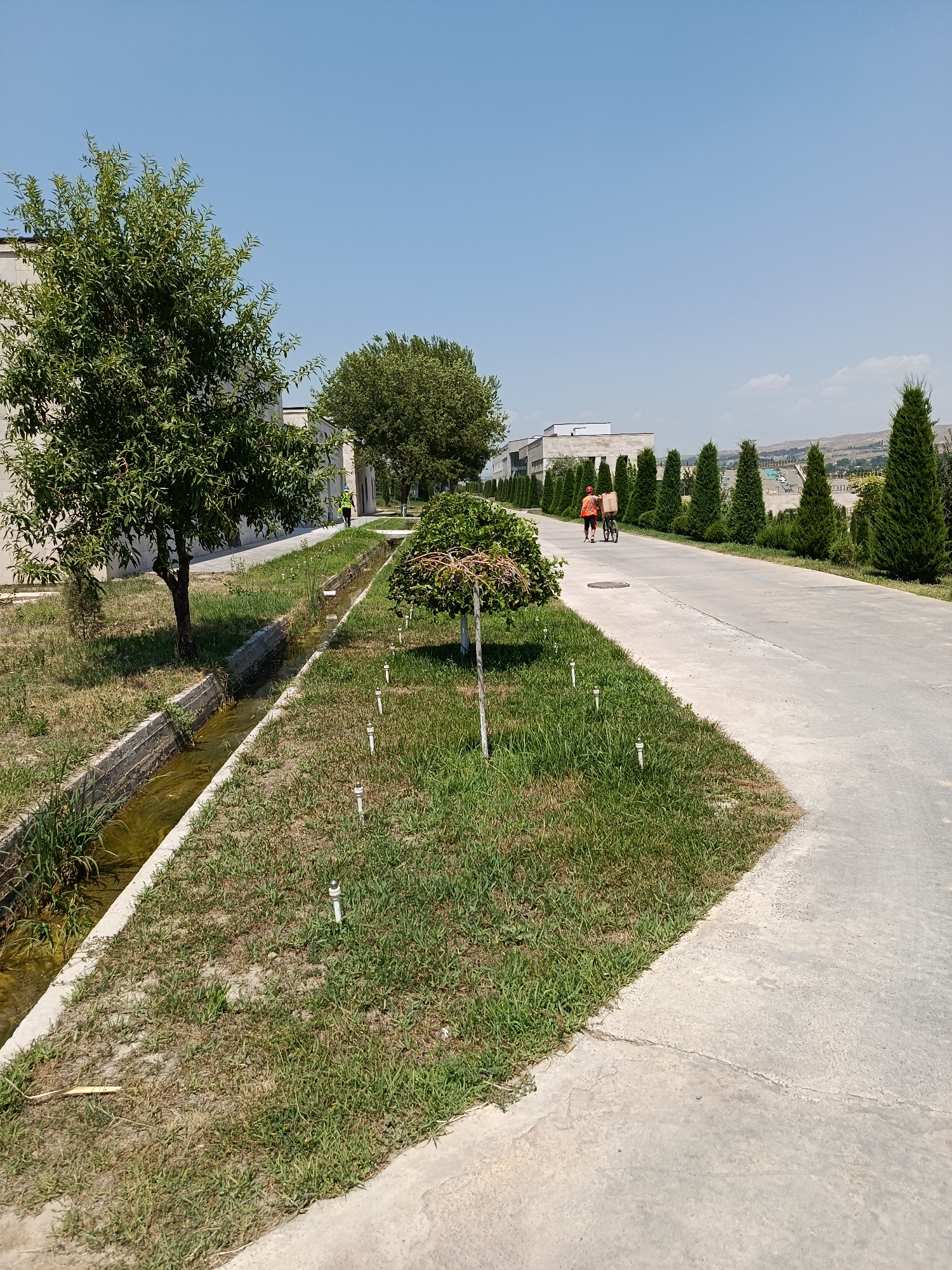 This is a medium shot, eye-level, outdoor, daytime, clear sky photograph taken in Gulshan, Uzbekistan. The scene depicts a paved walkway running along a grassy area bordered by a concrete irrigation ditch. On the left side of the walkway, there is a tree and a building. A person wearing a yellow vest is walking near the building. Further down the walkway, two people are seen from behind, one pushing a bicycle with a box on it. The walkway is lined with numerous small white posts, possibly part of a sprinkler system. In the background, there are more trees, buildings, and distant hills. The overall impression is one of a well-maintained, possibly institutional or residential, area in a warm climate.