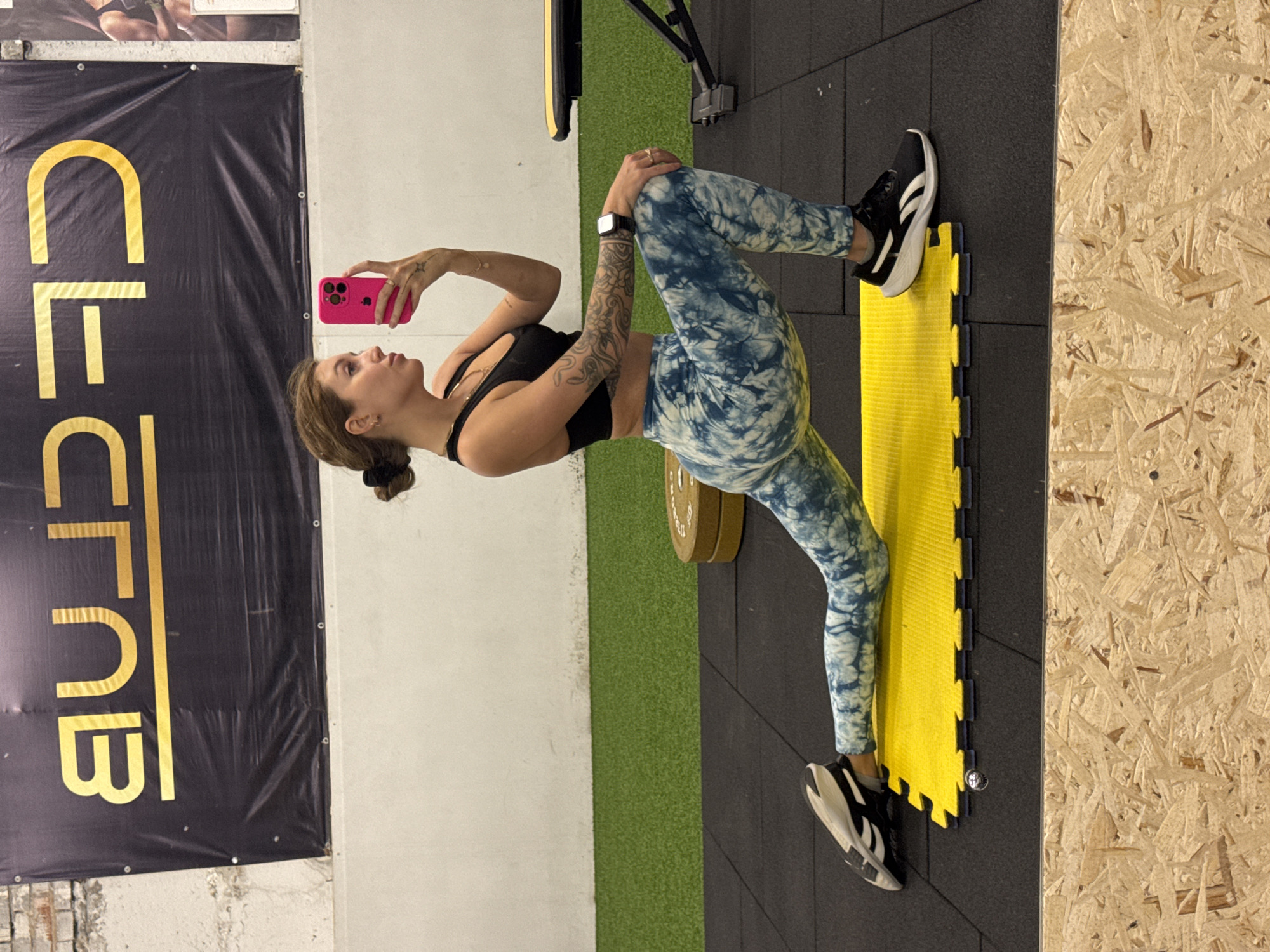 The image captures a young woman in an indoor gym setting, likely in Chaikovsky, Russia. She is lying on her back with her knees bent, positioned on a bright yellow interlocking foam mat that rests on black rubber gym flooring.
The woman has brown hair tied back and is wearing a black sports bra, blue and white tie-dye patterned leggings, and black and white athletic shoes. A prominent tattoo covers her left forearm, and a smaller star-shaped tattoo is visible on her right hand, where she also wears a ring. A black digital watch is on her left wrist. She holds a vibrant pink smartphone in her right hand, pointed upwards, suggesting she is taking a photo or video of herself in a pose, possibly related to exercise or fitness content creation. Her expression is focused and neutral.
The background reveals further details of the fitness environment. A large black banner with golden letters hangs on the wall behind her, clearly displaying the text "FLEX CLUB". To the right, a wall paneled with oriented strand board (OSB) provides a raw, industrial aesthetic. A strip of artificial green turf is also visible on the floor, and a weight plate can be seen partially on the floor near her midsection. The lighting is bright and consistent, indicative of an indoor facility.