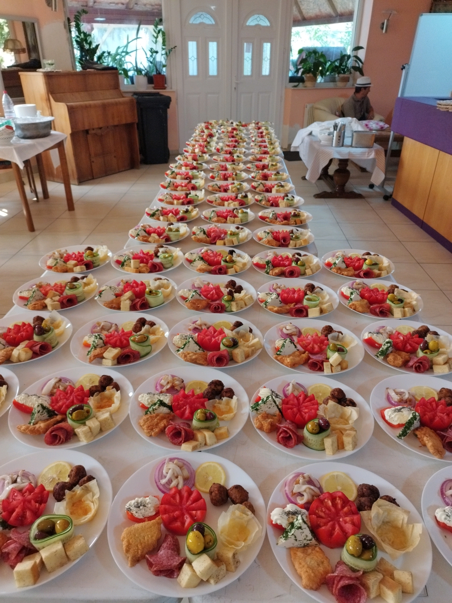 An outdoor event catering setup features rows of oval white plates, each meticulously arranged with appetizers. The plates are laid out on white tablecloths, creating long tables that extend into the background. The appetizers on each plate are consistent, consisting of cubed cheese, sliced tomatoes carved into flower shapes, rolled salami, olives, and small pastries. In the background, a man wearing a white hat and brown shirt sits on a chair near a table covered with a white tablecloth. There are also plants visible in pots and a large wooden cabinet. The overall scene suggests a prepared meal service, possibly for a celebration or gathering.