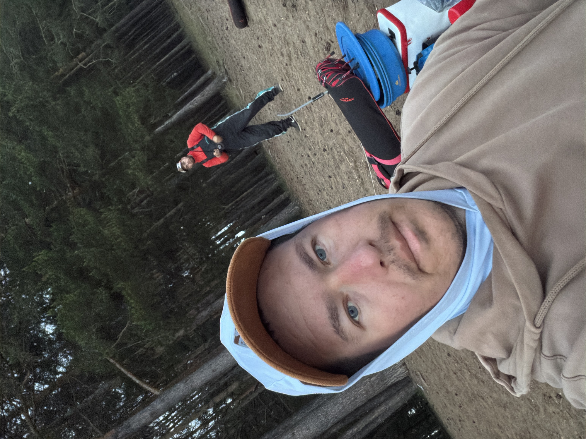 The image shows a selfie of a person with blue eyes, wearing a light blue hooded shirt and a light brown hooded jacket. To the left and slightly behind, another person is lying on the ground wearing a red and black jacket, dark pants, and a white helmet. They have their arms crossed over their chest and appear to be looking at the camera. Ski equipment including skis, ski poles, and a ski bag are visible nearby. The background features trees suggesting a wooded area. The setting appears to be on a dirt or sandy surface. The exact location in the city of Chaikovsky, Russia, is unknown.