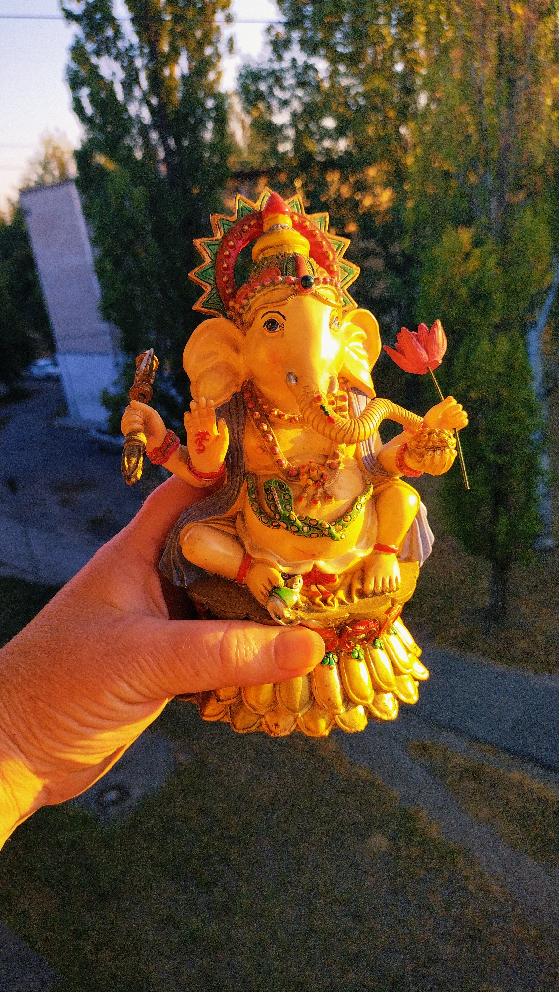 An extreme close-up shot captures a hand holding a golden statue of the Hindu deity Ganesha, illuminated by warm, low-angle sunlight. The statue, intricately detailed with jewelry and a headdress, is positioned in the foreground against a blurred background of trees, a building, and a paved area suggestive of an urban environment, likely in Horishni Plavni, Ukraine, given the context. The statue is adorned with beads, a green necklace, and a red flower in its upper right hand. The hand holding the statue is positioned to the left of the frame, with its fingers supporting the base of the figurine. The overall lighting suggests the photograph was taken during golden hour.