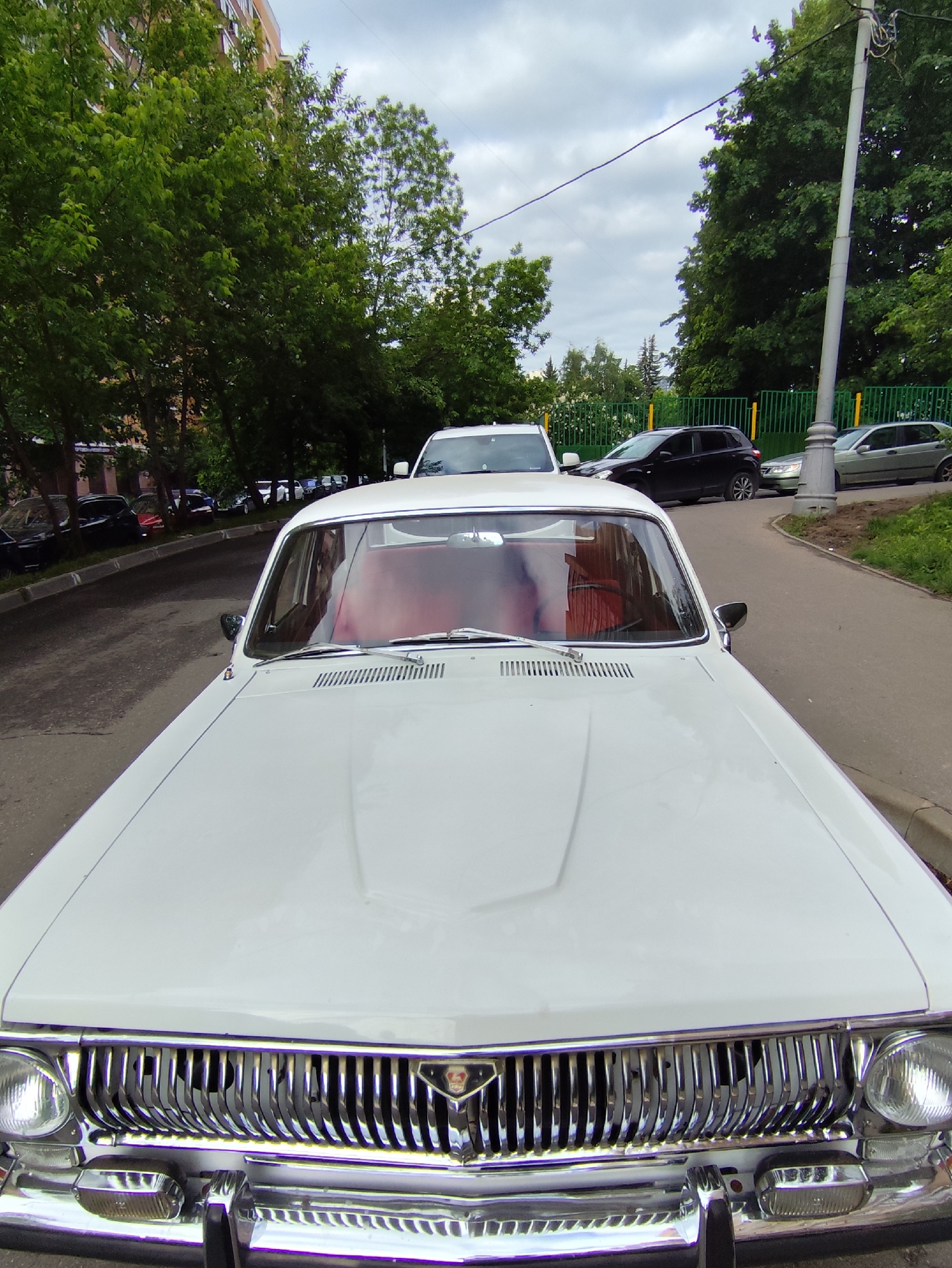The image shows a close-up of a white GAZ-24 Volga car parked on a street in Moscow, Russia. The car's front grille and hood are prominent, with the GAZ emblem visible. The interior appears to have red upholstery. In the background, other parked cars and a green fence can be seen, as well as trees and a building. The sky is overcast. The location appears to be a residential area with typical Moscow urban infrastructure.