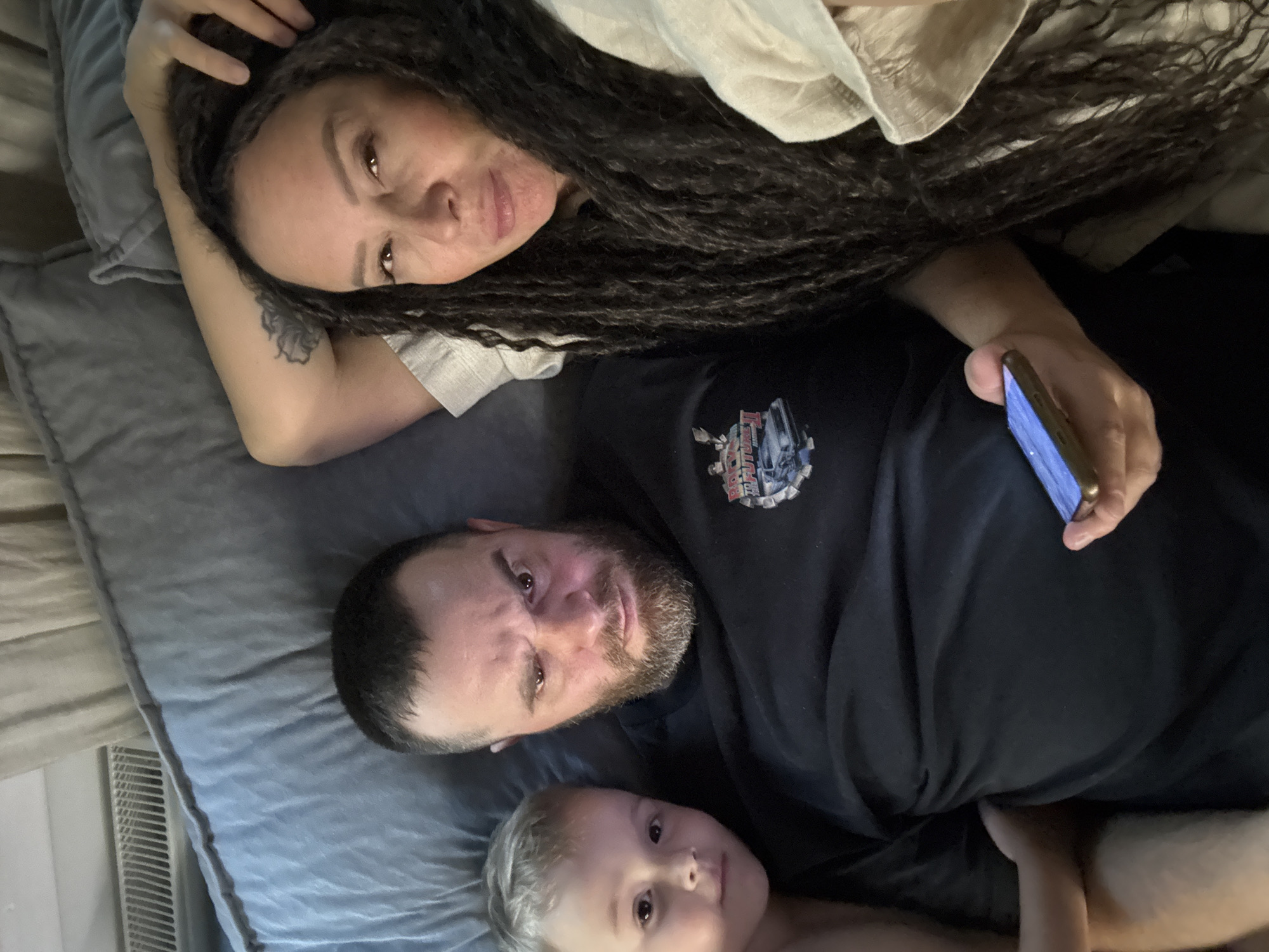 This indoor portrait, taken in Chaikovsky, Russia, features three individuals—a woman, a man, and a young child—lying together on what appears to be a bed or sofa, covered by a grey, subtly patterned blanket or comforter. The lighting suggests an ambient indoor setting.
At the top of the frame, a woman with long, dark, curly hair and brown eyes lies on her back, her head resting on a pillow. A tattoo is visible on her upper left arm. She holds a smartphone with a bright screen in her right hand, looking towards the camera with a neutral to slightly smiling expression.
Below her, a man with a beard and short dark hair is also lying on his back. He wears a black t-shirt featuring a graphic print of the DeLorean time machine from the movie "Back to the Future." Visible text on the shirt reads "BACK TO THE FUTURE" and "PART III." He looks directly at the camera with a calm expression.
At the bottom, a young child with light-colored hair is nestled beside the man, also looking towards the camera with a soft, curious gaze. On the far left, a white radiator or vent is partially visible, suggesting an interior wall. The overall scene conveys a relaxed and familial atmosphere.