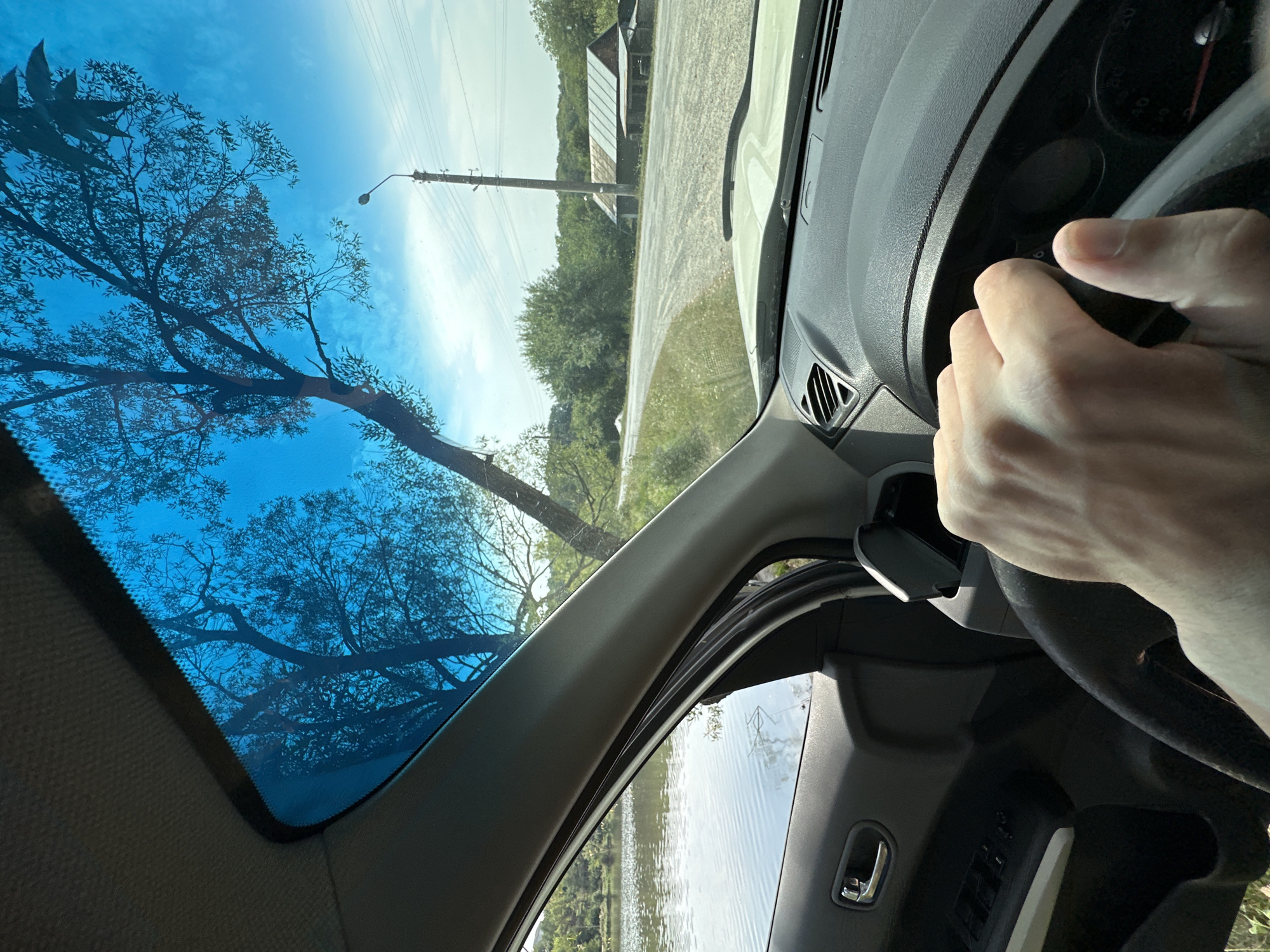 The image is taken from inside a car, looking through the windshield. A hand is visible gripping the steering wheel. Outside the car, a body of water is on the left, and a road with trees, a utility pole, power lines, and two rustic buildings are visible in the distance on the right. The sky is blue with some clouds. The scene appears to be a rural or semi-rural area.
