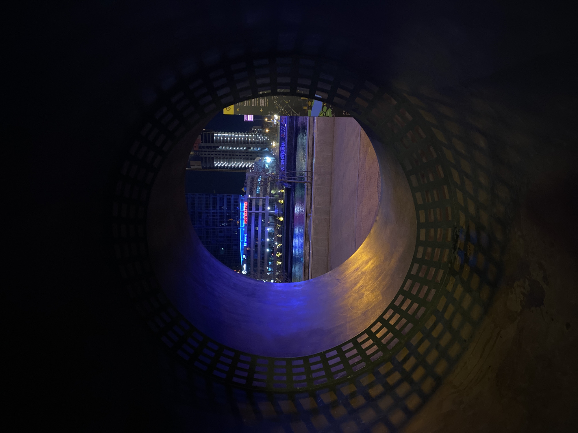 The photograph, taken at night, depicts an urban cityscape as seen through a large, circular aperture. The aperture appears to be part of a pipe or tunnel structure. The city visible through the opening is identified as Yekaterinburg, Russia, based on the presence of the "Polototoptran" building sign. The aperture casts intricate shadows on its interior surface. Outside, buildings are illuminated with a mix of white, blue, and yellow lights. The scene presents a juxtaposition of the confined, artificial structure of the aperture with the vibrant, dynamic cityscape beyond.