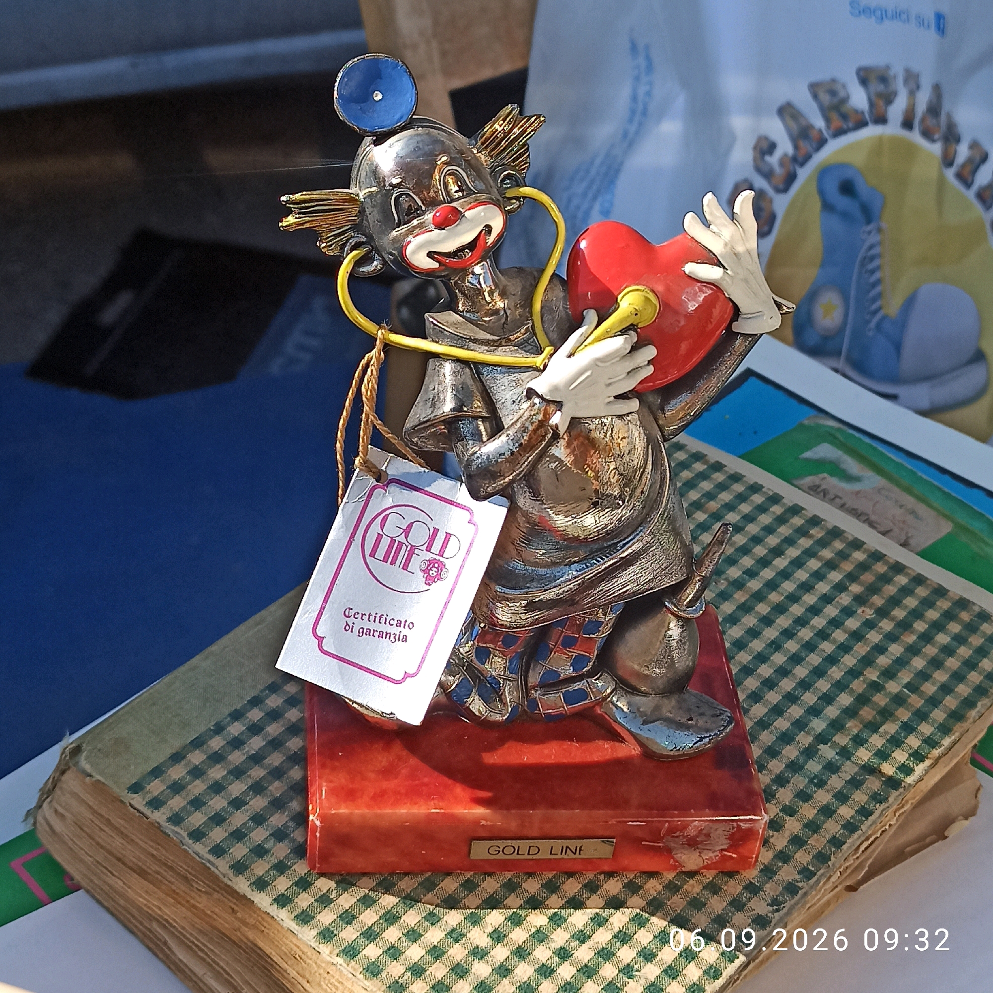 A metallic clown figurine, adorned with a blue stethoscope and holding a red heart, is displayed on top of a book with a green and white checkered cover. The clown wears white gloves and has a painted red nose and smile. A white tag with the "GOLD LINE" logo and "Certificato di garanzia" (Guarantee Certificate) is attached to the figurine. The figurine is mounted on a red base with a "GOLD LINE" label. In the background, a white bag with Italian text and a picture of blue boxing gloves is visible. The scene appears to be set outdoors, possibly at a market or fair, with a glimpse of a blue surface on the left and a sunlit area on the right. A date and time stamp "06.09.2026 09:32" is present in the lower right corner, suggesting this image was taken on that date and time. The overall impression is of a commercial or display setting, possibly in Sannicandro di Bari, Italy, given the Italian text on the tag and background bag.