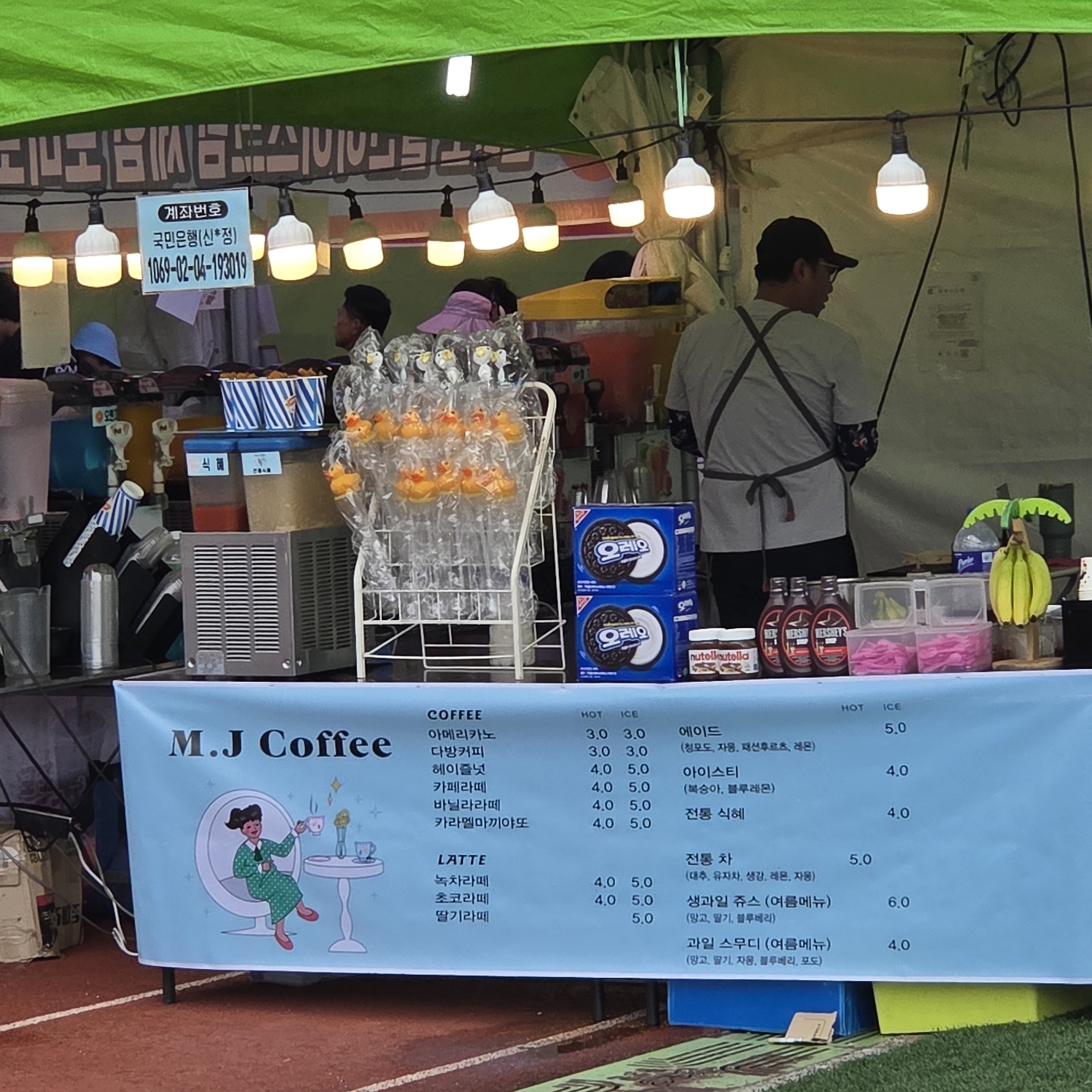 The image shows an outdoor M.J. Coffee stand in Dunnae-myeon, South Korea. The stand is set up with a green canopy, string lights, and various beverage dispensers. A menu board lists coffee, latte, and other drink options with prices. Among the displayed items are Oreo cookies and jars of Nutella. In the foreground, a blue banner features the M.J. Coffee logo with an illustration of a woman enjoying coffee. A man wearing an apron is visible behind the counter, appearing to serve customers. The background suggests a public outdoor setting, possibly a park or market, with other people and tents.