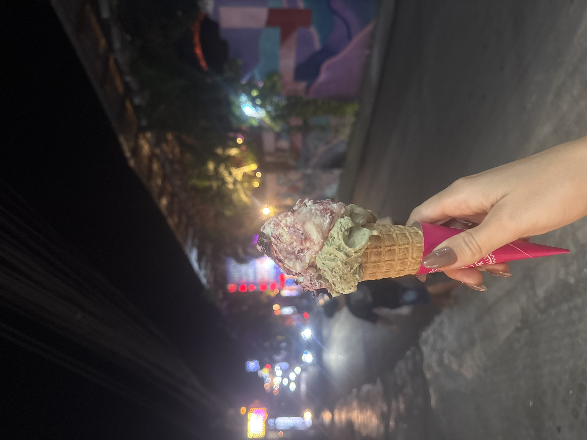 A person's hand with long, manicured nails holds a pink-wrapped ice cream cone with two scoops of ice cream in what appears to be Kuta Selatan, Indonesia at night. The ice cream cone has a waffle pattern. The top scoop is a lighter color with visible chunks of fruit or candy, and the bottom scoop is a pale green. The background is blurred and illuminated by streetlights and colorful building lights, suggesting an urban setting. A street with cars and a person walking is visible in the distance. The overall scene depicts a casual evening outing.