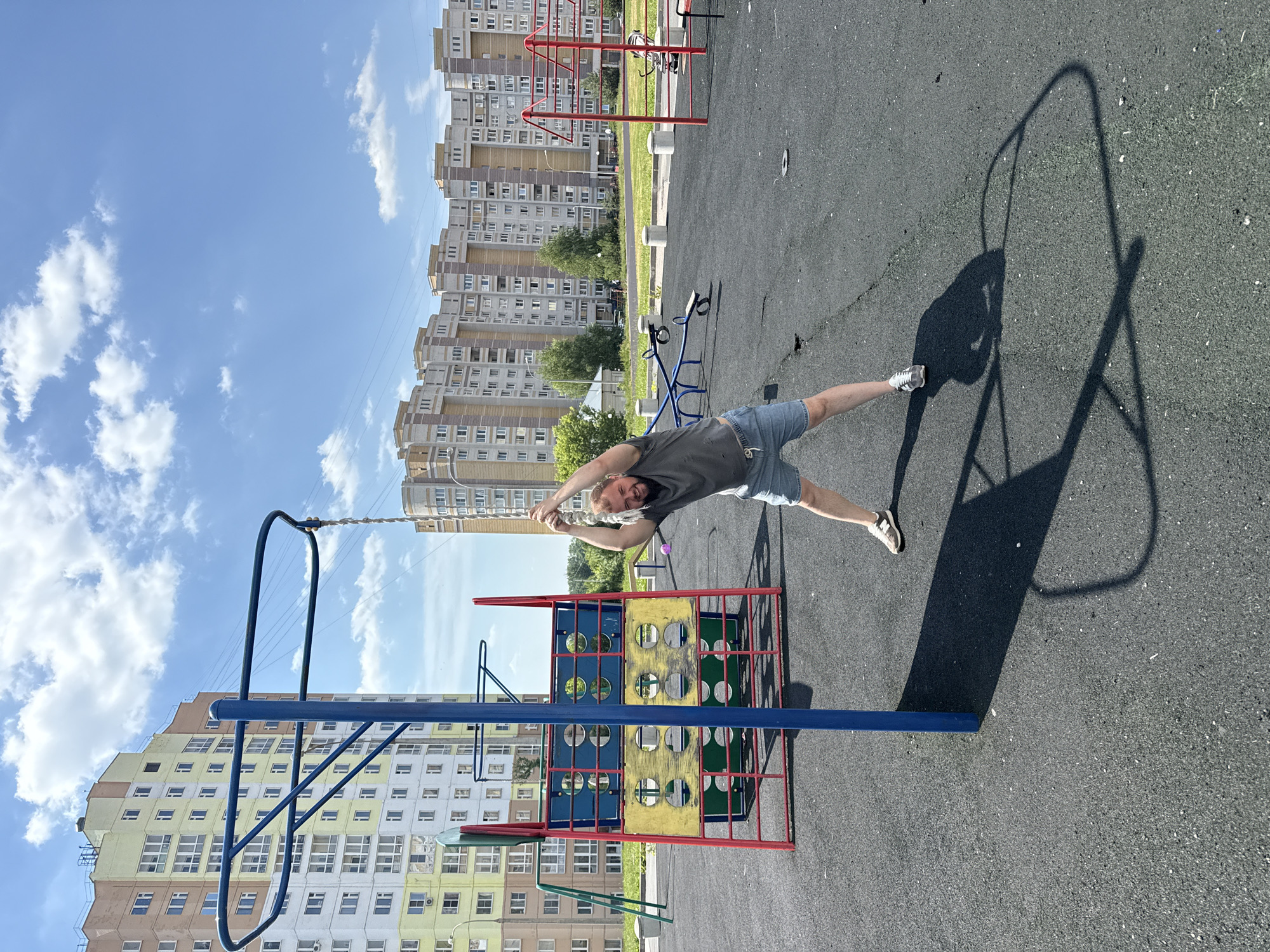 A man with a beard, wearing a gray t-shirt and denim shorts, is on a playground. He is holding onto a rope hanging from a blue and red structure, with his arms outstretched and body in a bent position, seemingly doing some form of exercise or play. The playground has a black rubberized surface. In the background, there are several tall apartment buildings, characteristic of modern urban development, with a clear blue sky and scattered white clouds above. The architecture of the buildings, particularly the color palette and design elements like the balconies and windows, suggests an Eastern European context. The presence of multi-story residential buildings in the background and the overall urban setting indicate this is a playground located within a residential district of a city. Given the architectural style and the specific building designs with their color blocks, this scene is consistent with residential areas found in the city of Vladimir, Russia.