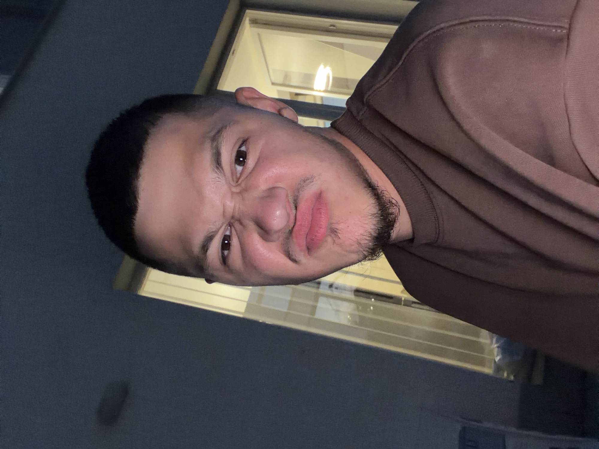 A young man is captured in a close-up, selfie-style shot, making a displeased facial expression. He has dark, closely cropped hair and a light beard. He is wearing a brown sweatshirt. Behind him, a large window reveals blinds and interior lighting, suggesting the photograph was taken indoors at night, possibly in a residential setting. The background on the left is dark and indistinct.