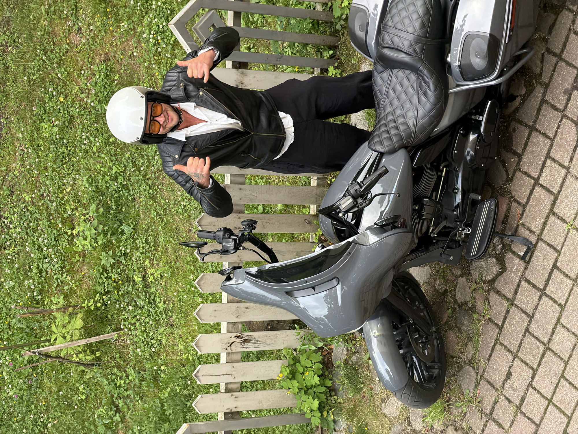 A man wearing a white helmet, orange sunglasses, a black leather jacket, and a white unbuttoned shirt stands next to a grey motorcycle. He is giving a thumbs-up gesture with both hands. The motorcycle is parked on a cobblestone pathway in front of a white picket fence. Behind the fence is a grassy area with some small white flowers and a wooden stake. The scene appears to be in a residential outdoor setting.