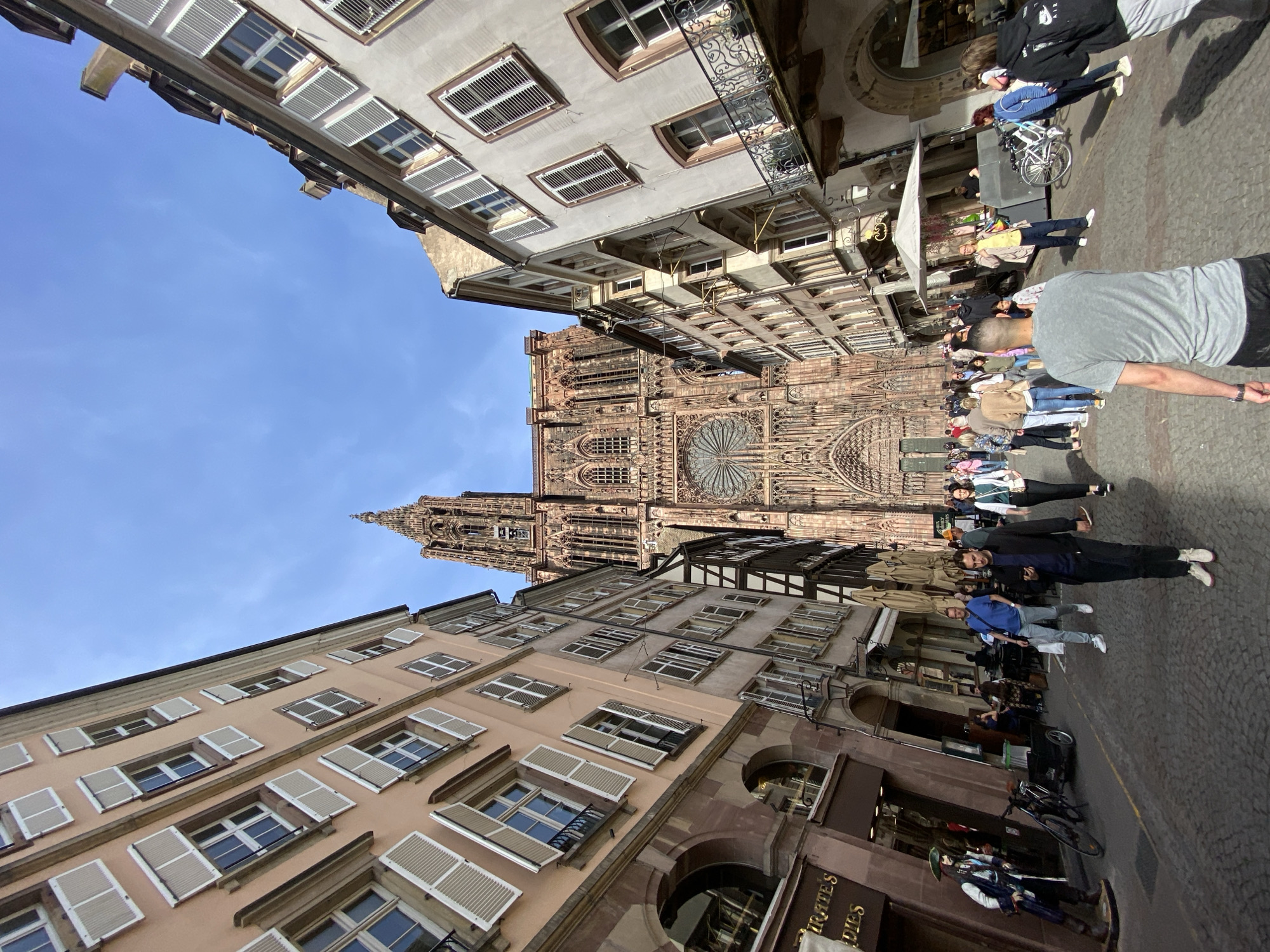 The image, taken in Strasbourg, France, captures a bustling street scene dominated by the majestic Strasbourg Cathedral in the background. The photograph is taken from a low angle, looking up towards the cathedral and the surrounding buildings.
The central subject is the grand Gothic cathedral, showcasing intricate stone carvings, a large rose window, and a towering spire on its right side. On either side of the street, multi-story historical buildings line the view. These buildings feature numerous windows, many adorned with open or partially closed white louvered shutters. Some structures exhibit traditional half-timbered elements, adding to the historic charm.
Numerous people are visible on the cobblestone street, engaged in casual activities like walking and sightseeing. They are dressed in everyday clothing, including t-shirts, hoodies, and jeans. Some individuals are looking towards the cathedral, while others walk past shops and cafes. A white tent or canopy is set up on the right side of the street, possibly for an outdoor vendor or seating area. Bicycles are parked in various spots.
The scene is illuminated by bright daylight, casting clear shadows and indicating a sunny day with a clear blue sky. A notable object in the foreground, on the right, is a detailed pirate statue standing outside a shop. Visible text on the shop sign reads "PIRATES" and "DES", suggesting a pirate-themed establishment. Another partial sign may read "VOYAGES" or "HOTES". The overall atmosphere appears lively and relaxed.