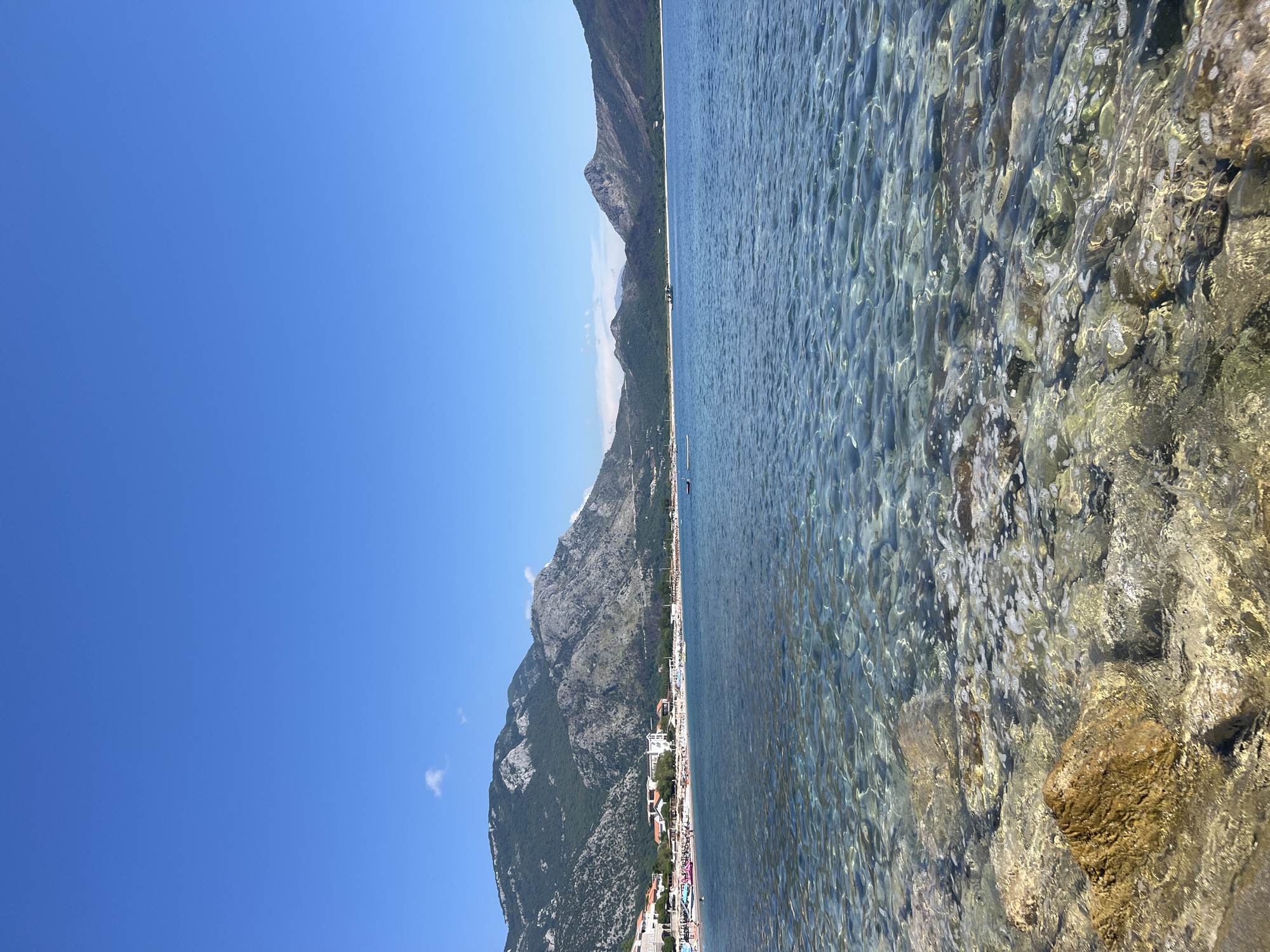 This is a landscape image taken from a low angle, looking out over a body of water towards a coastline and mountains. The right side of the image is dominated by clear, shallow water with visible rocks and pebbles on the seabed. The water is disturbed by gentle ripples, creating a textured surface.
The left side of the image shows a shoreline with buildings, indicating a populated area. Beyond the buildings, steep, rocky mountains rise dramatically, covered in sparse vegetation. The sky is a vibrant, cloudless blue, suggesting a clear and sunny day. A few faint clouds are visible near the mountain peaks.
The scene appears to be a coastal town or resort area. The presence of buildings along the beach and the clear water suggest a holiday or recreational setting. There are no clearly identifiable people or specific objects that stand out, other than the natural elements and the settlement. The overall mood is peaceful and idyllic, evoking a sense of a warm, sunny day at the beach. The image is oriented vertically, with the horizon line running diagonally across the frame. No visible text is present in the image.
