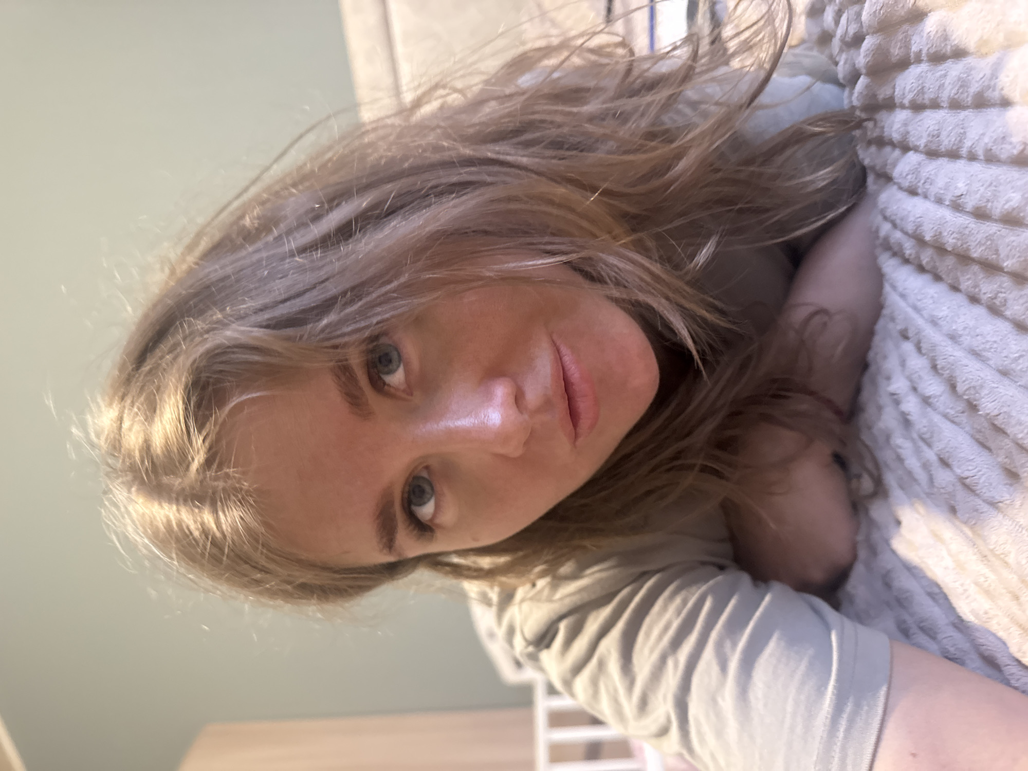 A woman with long, light brown hair and blue eyes is lying on a textured, light-colored blanket. She is wearing a light-colored shirt. The background includes a light-green wall and a partially visible light-wood furniture surface. A white structure, possibly part of a chair or other furniture, is visible in the lower center of the image. The location of Ферма, Russia is not discernible from the image.
