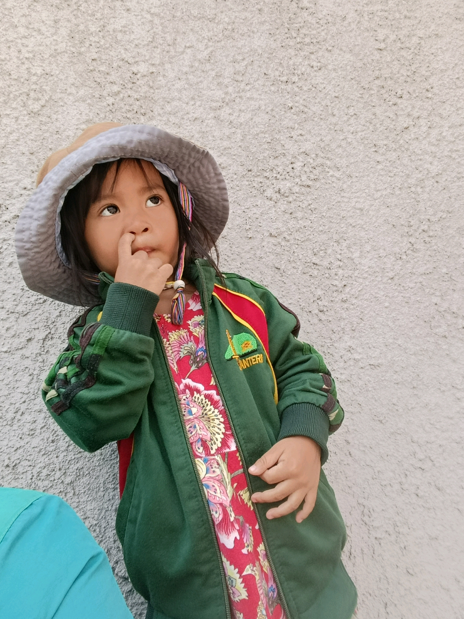 In Babelan, Indonesia, a young girl with dark hair and brown eyes stands against a textured light gray wall. She wears a light brown wide-brimmed hat with a gray lining, and a green jacket over a red floral print dress. Her right index finger is in her nose. A turquoise garment is partially visible to the left of the frame.