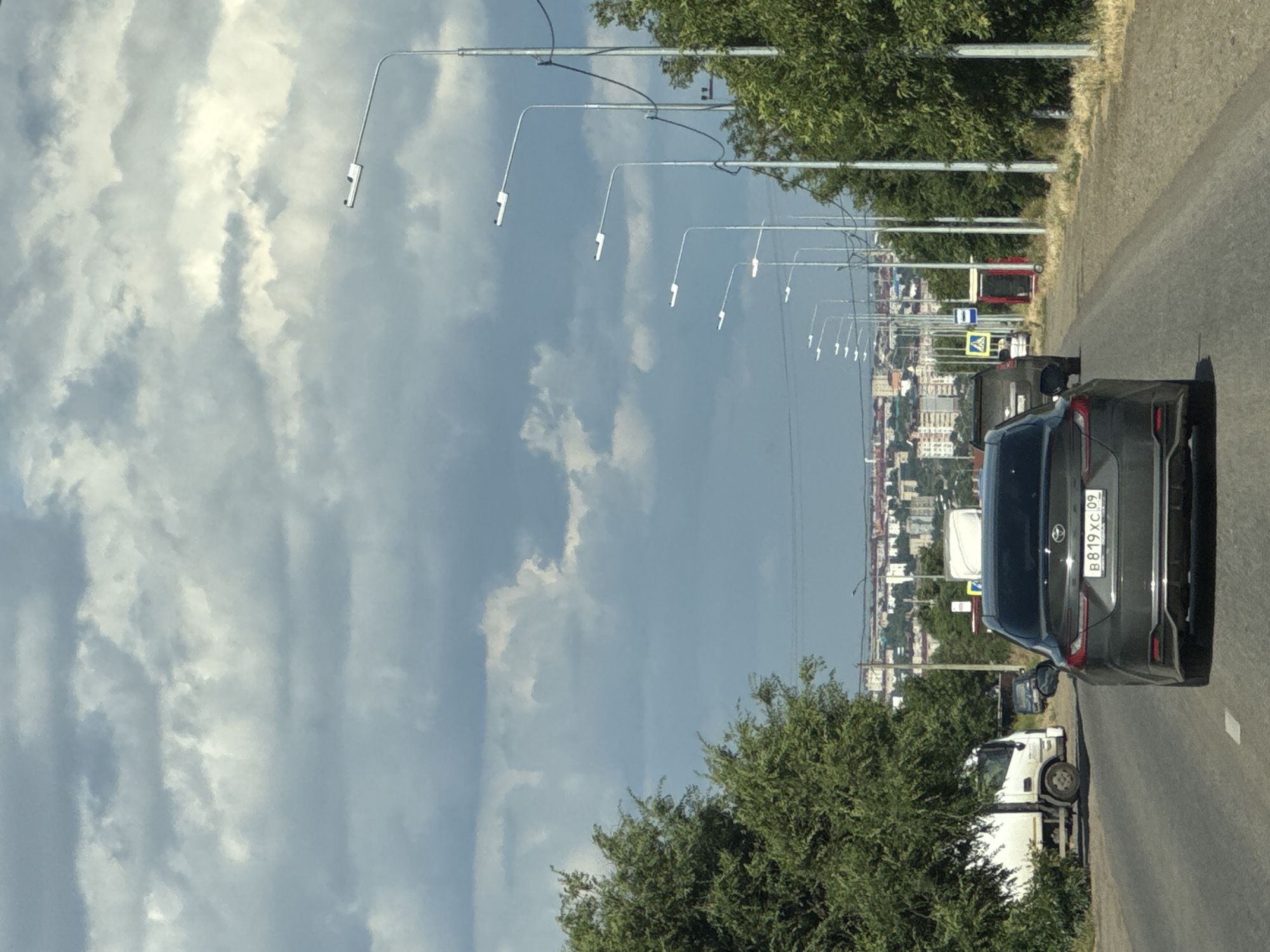 This image, taken roadside in Russia, depicts a cloudy day with a view of a cityscape in the distance. The foreground is dominated by a grey car with the license plate "B819XC 09" visible. To the left of the car, a white truck is parked. The road extends into the distance, lined with numerous streetlights that curve upwards and away from the viewer. Dense green trees line the right side of the road, bordering the asphalt. In the background, buildings of varying heights and colors form a town or city skyline. The sky is filled with dramatic, cumulus clouds, suggesting it might be midday or late afternoon. The overall scene conveys a sense of everyday travel along a suburban or semi-urban road.